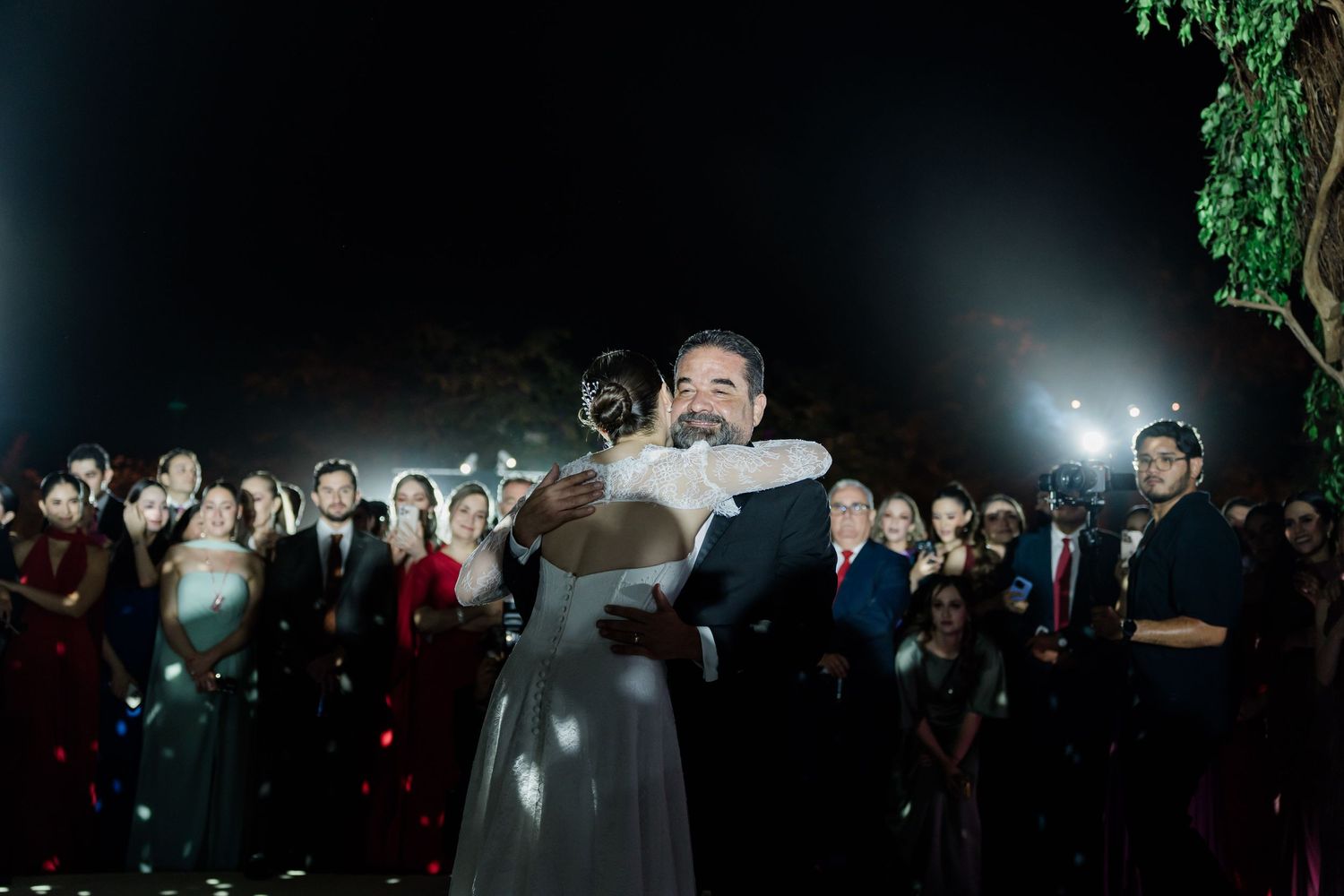 Bride and groom share a romantic first dance embrace surrounded by wedding guests at an outdoor nighttime reception.