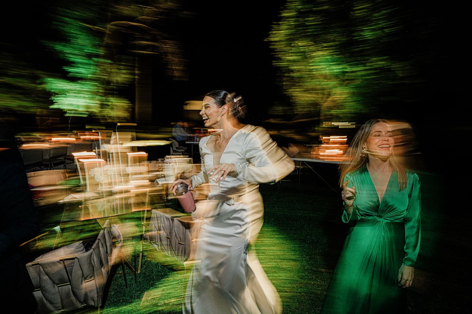 Bride in white dress and guest in green gown walking at illuminated outdoor wedding reception.
