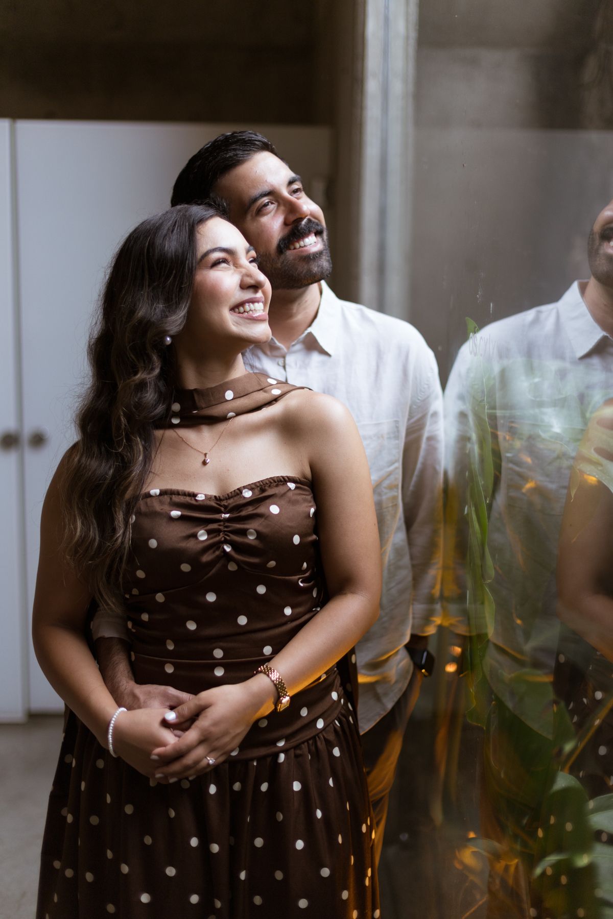 Smiling couple standing together near glass reflection, woman in brown polka dot dress and man in white shirt.