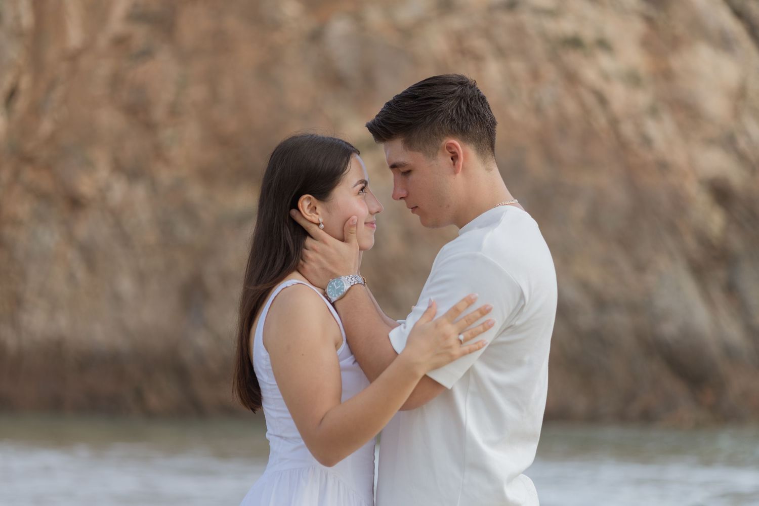 A young couple in white clothing share a romantic moment on a rocky beach, gazing into each other's eyes.
