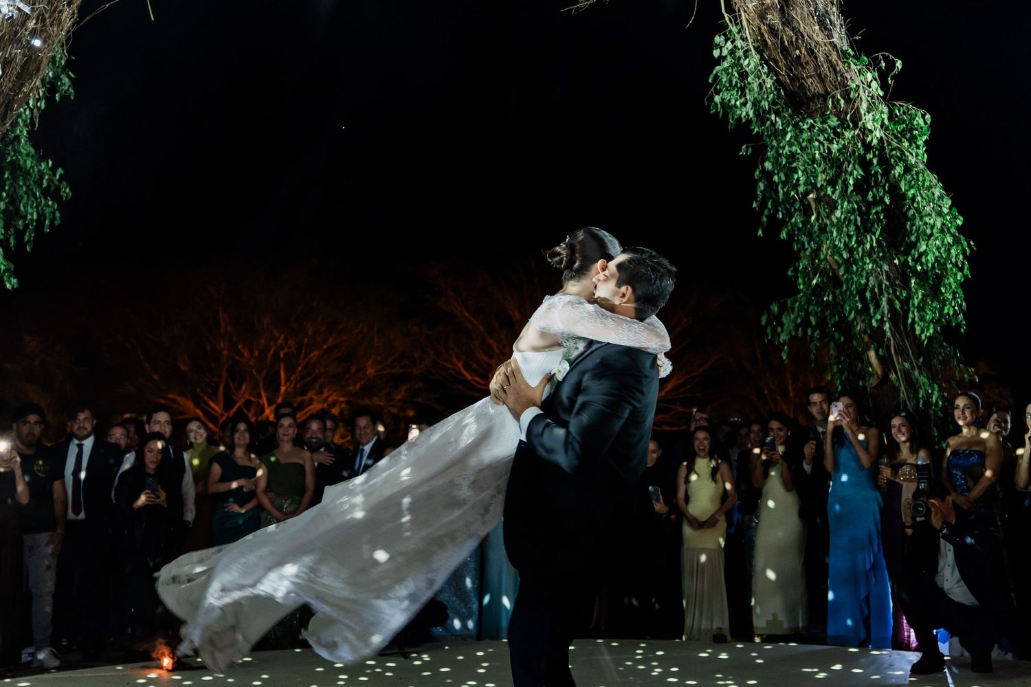 Groom lifts bride in a joyful first dance under a beautifully lit tree at an outdoor wedding reception.