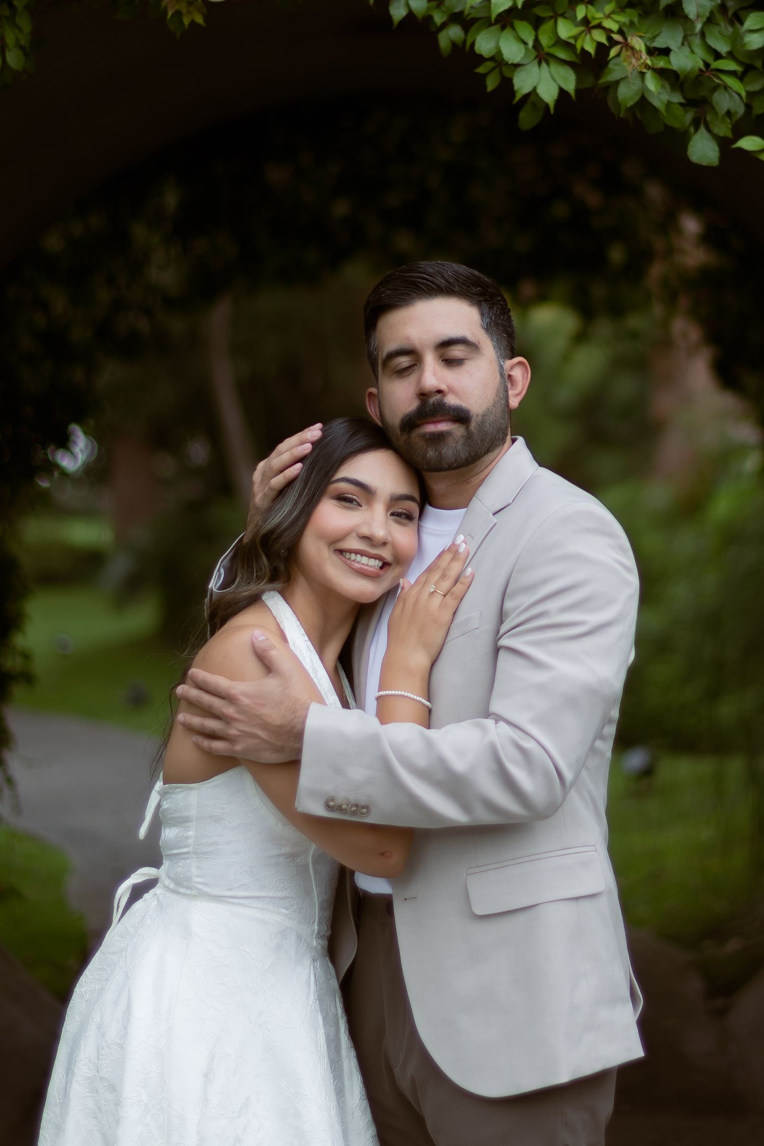 Groom in beige suit embraces smiling bride in white halter dress under lush green trees.