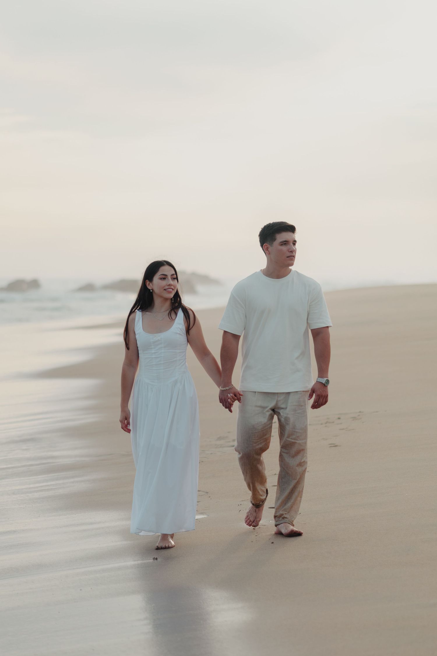 Couple holding hands walking barefoot on a sandy beach at sunset, woman in white dress, man in white shirt.