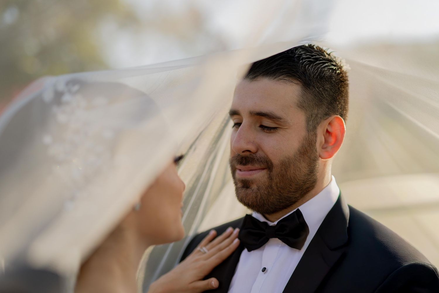Romantic close-up of groom smiling tenderly at bride beneath flowing wedding veil in soft light.