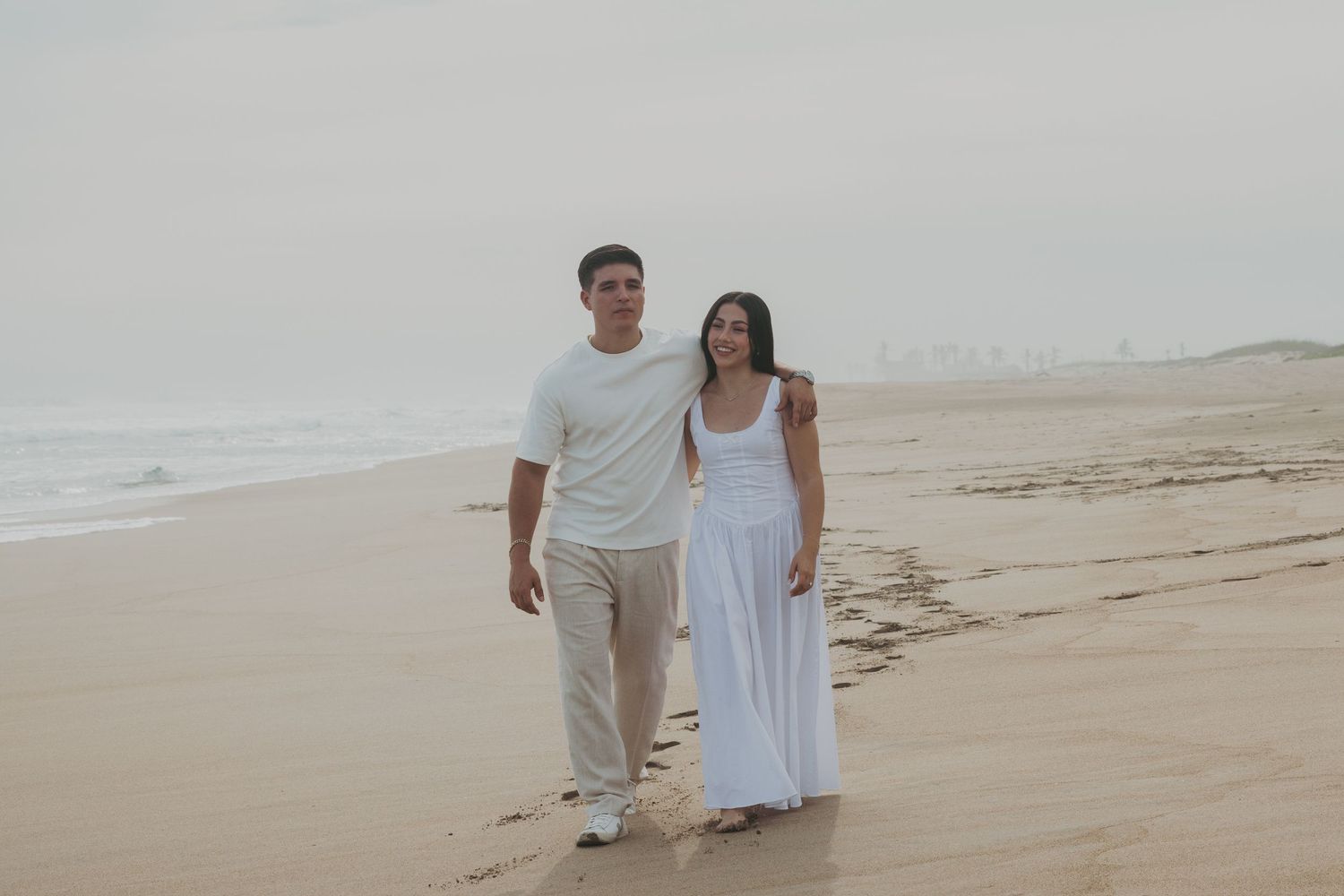 Couple walking hand in hand on a misty beach, wearing white outfits, with ocean waves in the background.