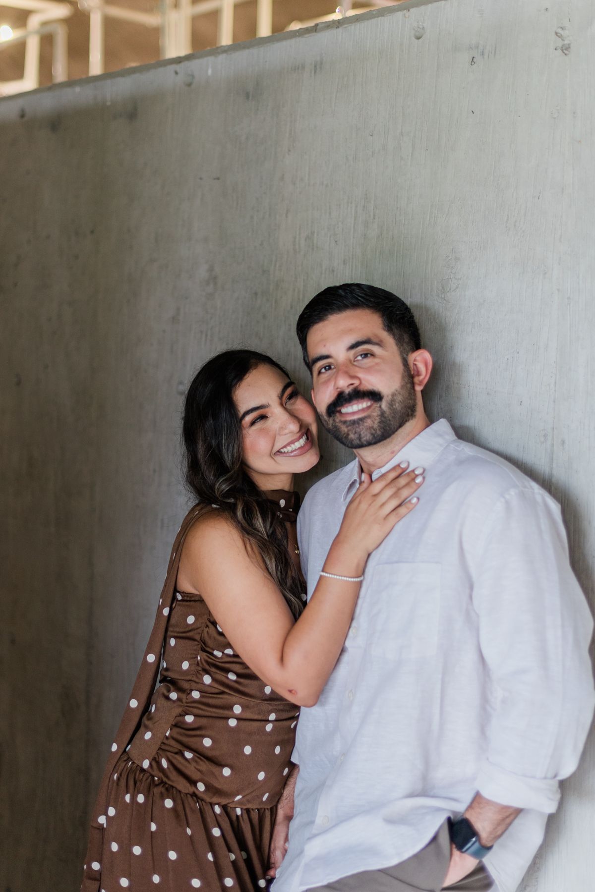 Joyful couple leaning against concrete wall, woman in brown polka dot dress embracing smiling bearded man.
