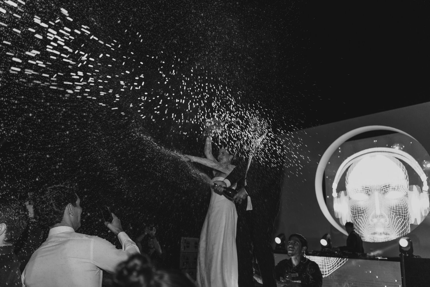 Black and white photo of bride spraying champagne at a festive wedding reception with DJ in background.