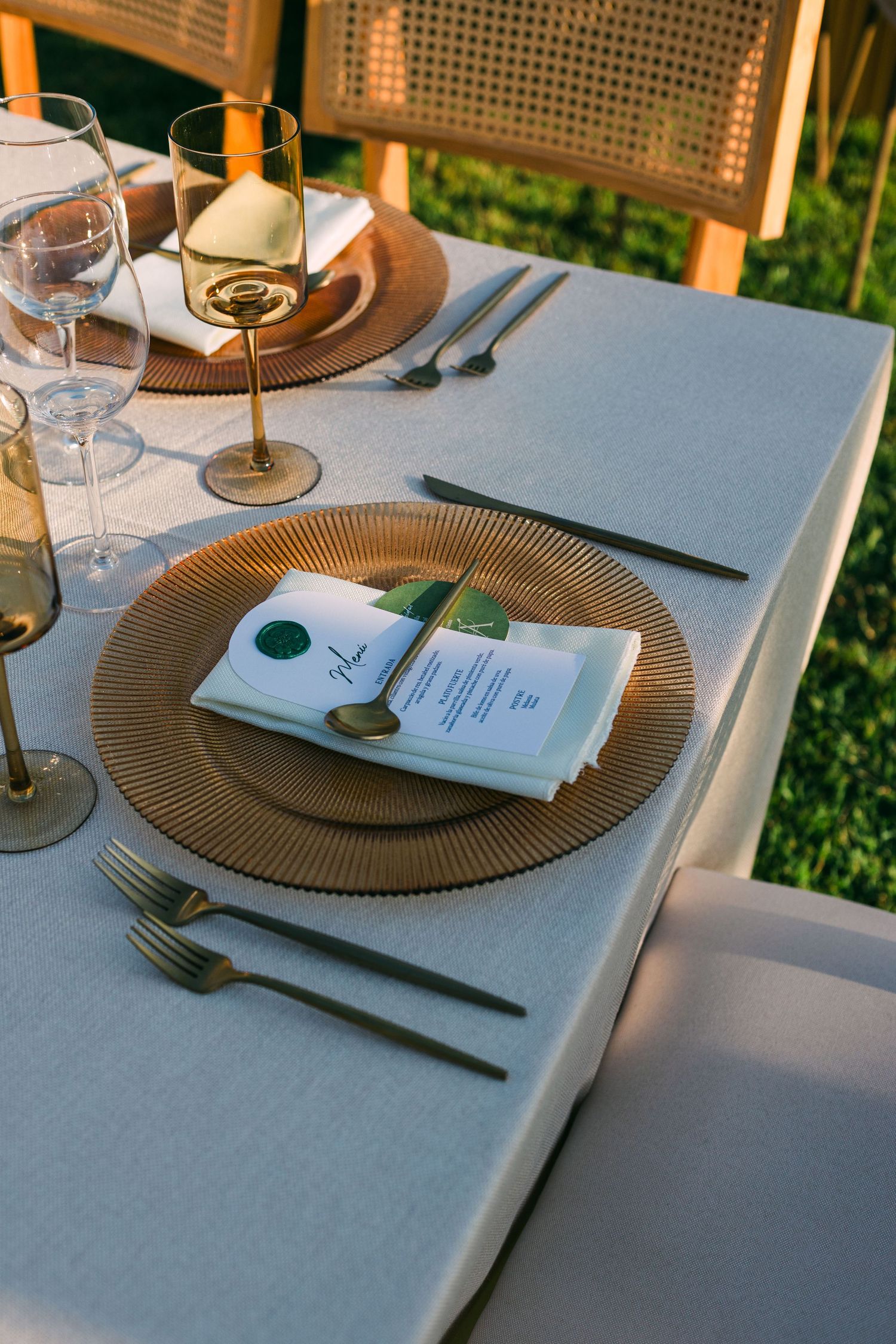 Gold charger plate with menu card, green wax seal, and dark gold cutlery on white linen tablecloth.