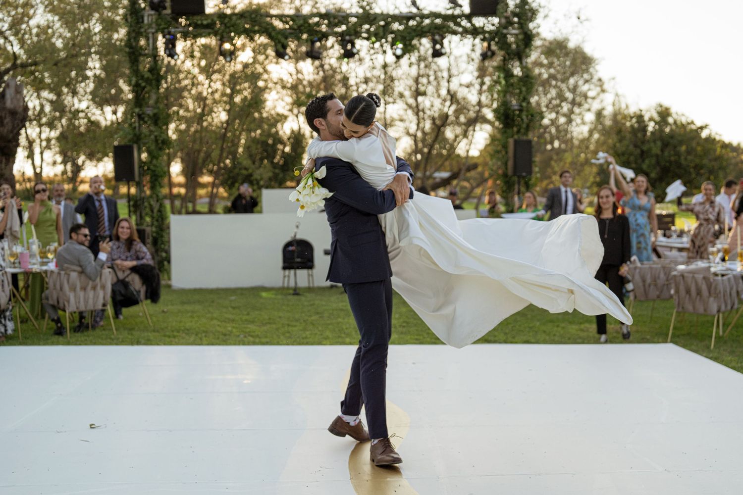 Groom lifts bride in white wedding dress during first dance at outdoor evening reception.