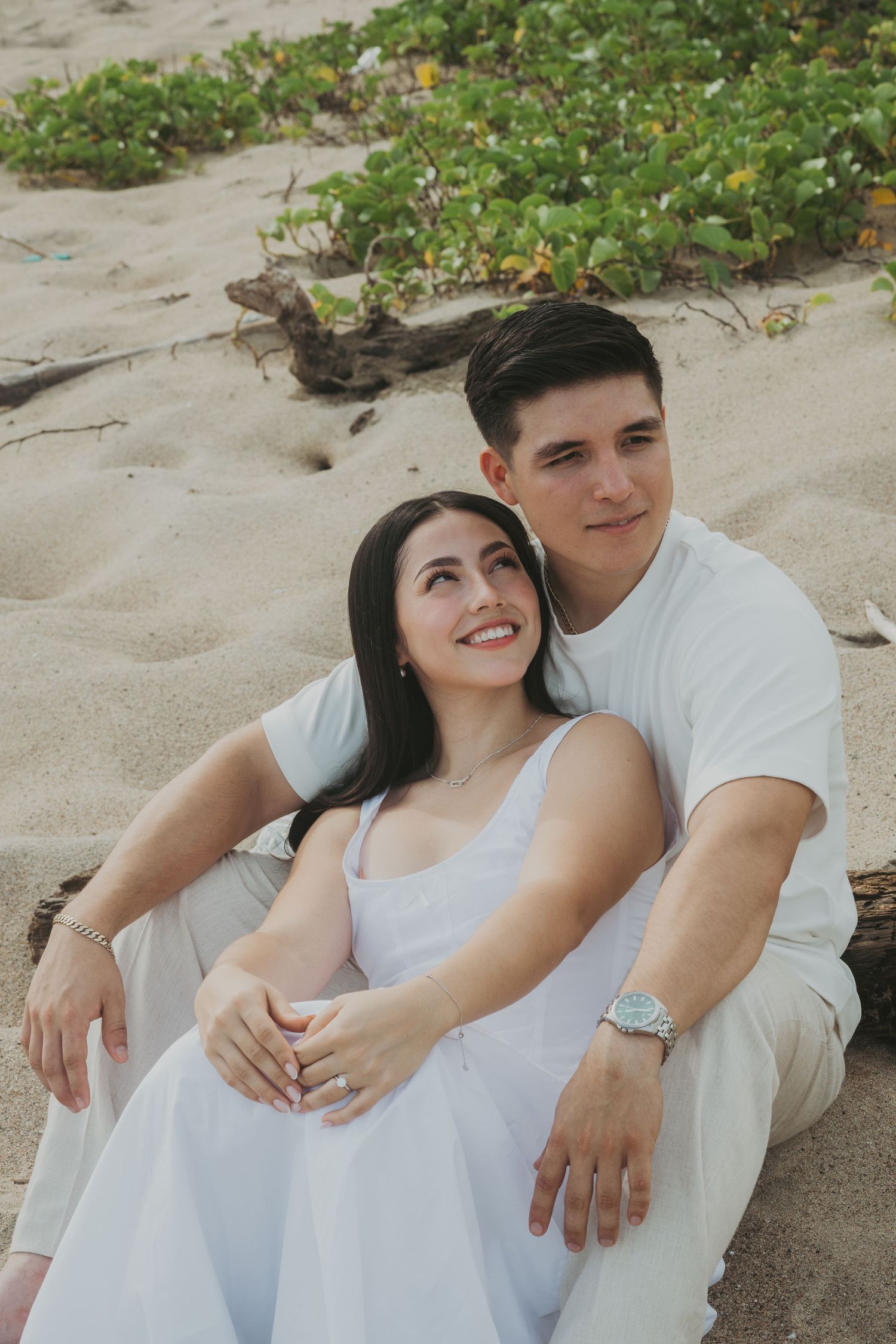 A smiling couple in white outfits sits together on a sandy beach, the woman leaning against the man romantically.