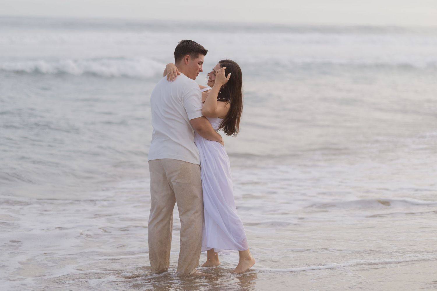 Romantic couple embracing on a sandy beach at sunset, wearing white outfits with ocean waves in the background.