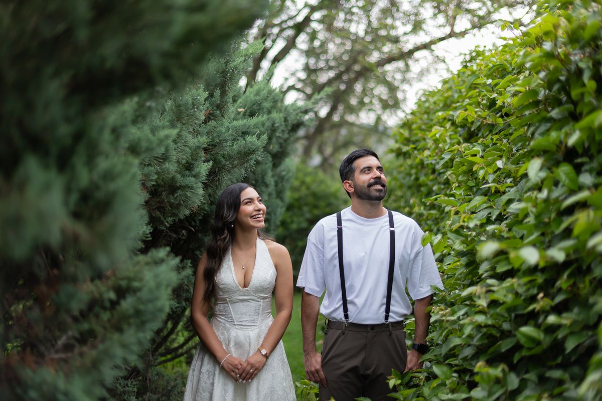 Smiling couple walks hand in hand through lush green garden path, woman in white dress, man in suspenders.