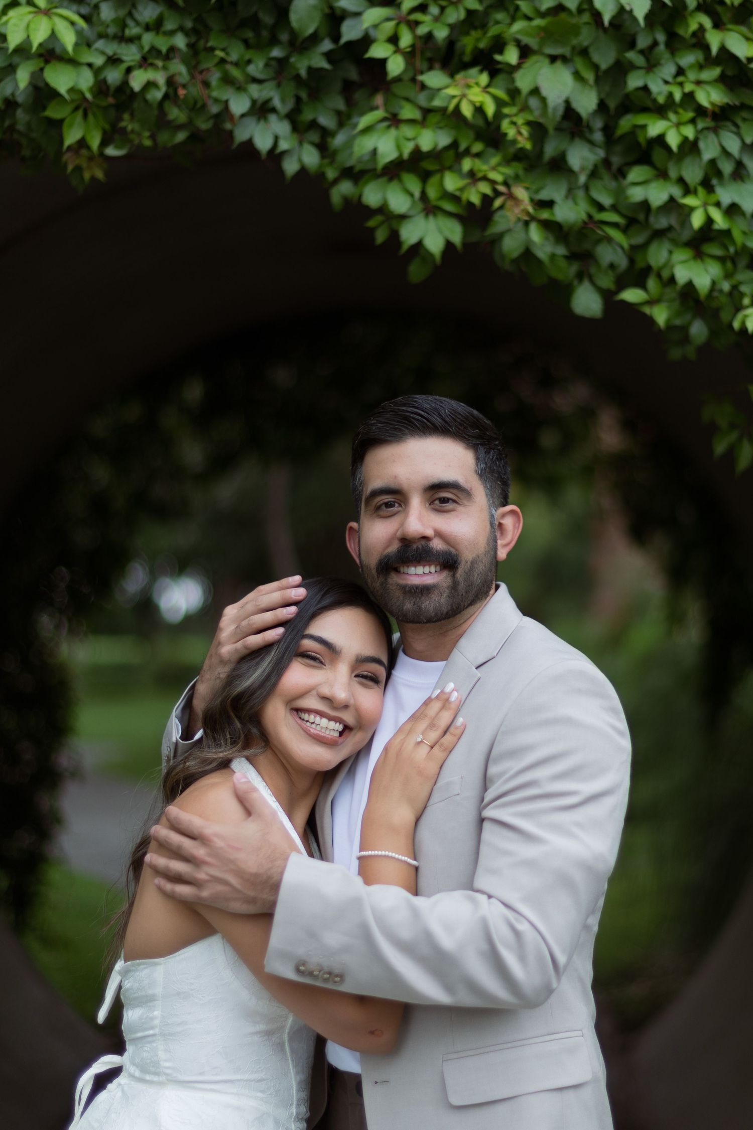 Smiling couple embraces under ivy-covered arch, bride in white dress and groom in beige suit.