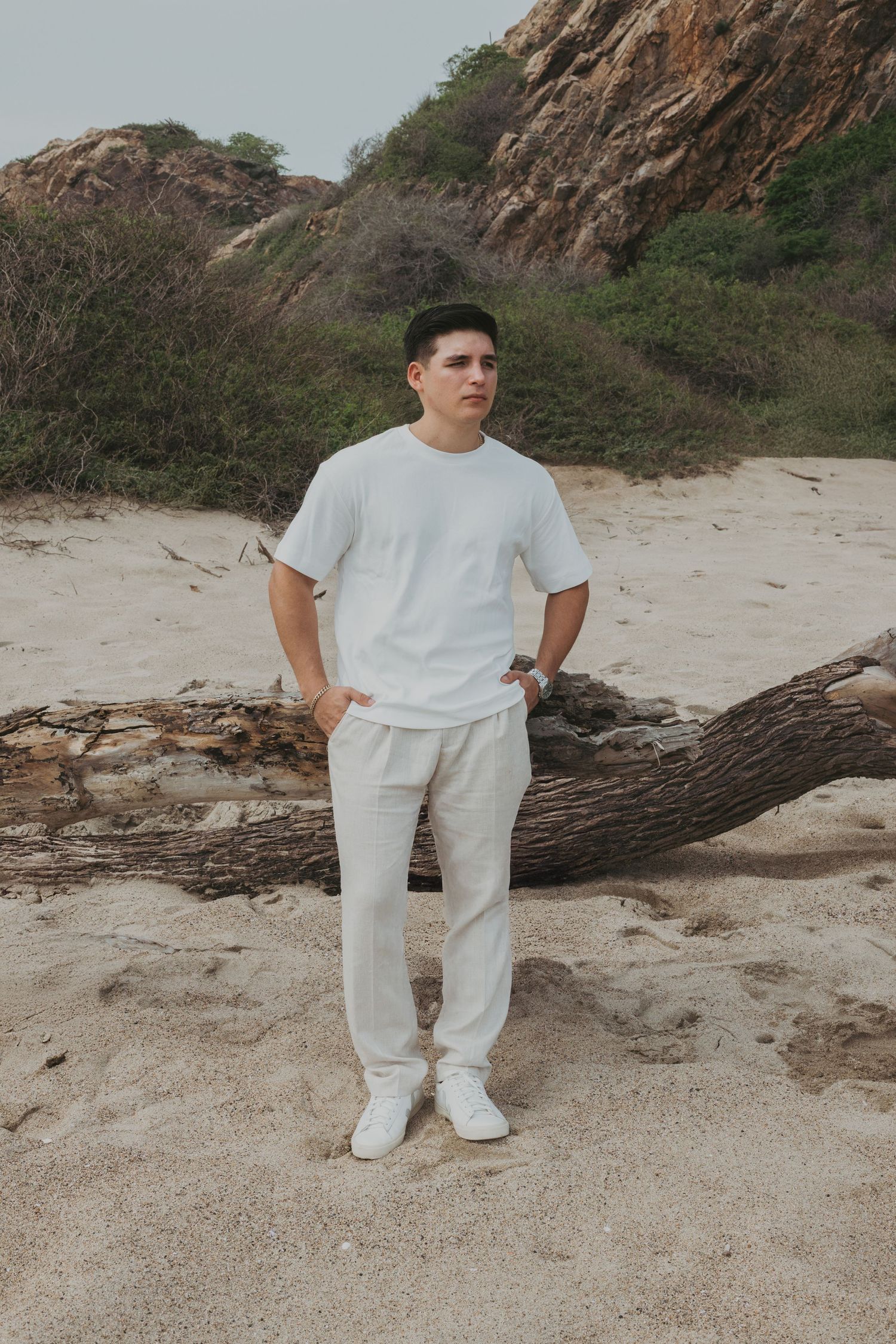 Young man in white outfit standing on sandy beach near driftwood with rocky cliffs and greenery behind.