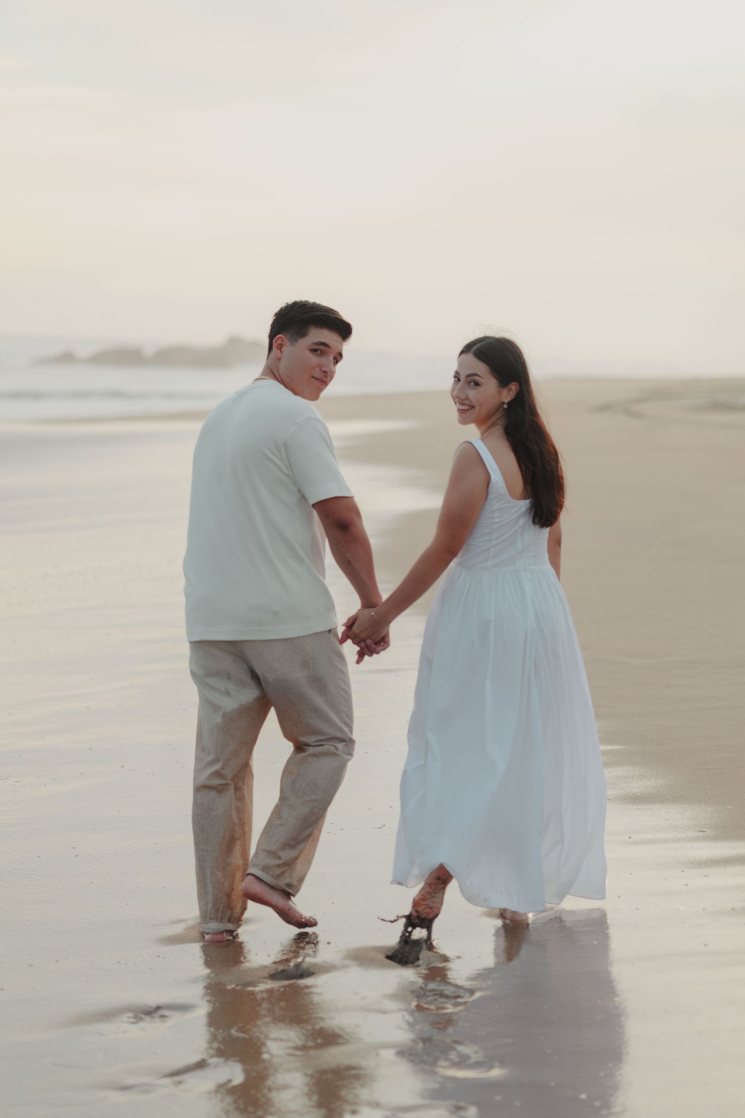 Couple holding hands walking on a beach at sunset, woman in white dress and man in casual attire.