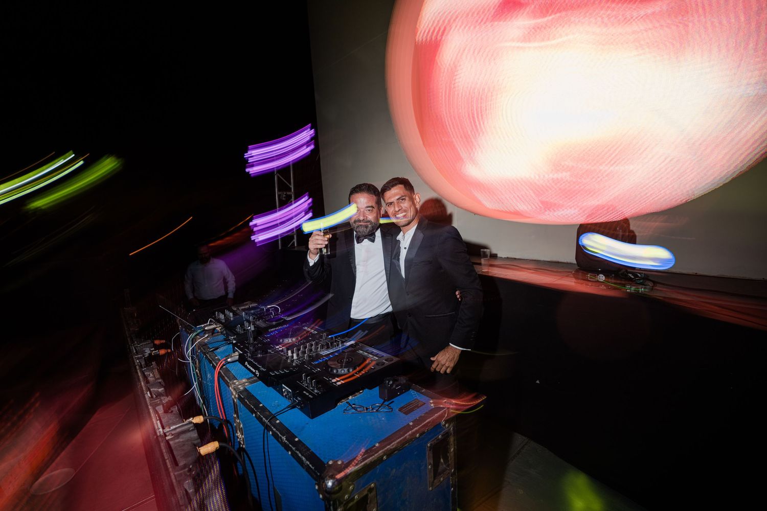 Two men in tuxedos posing at DJ booth with colorful lights at wedding reception.