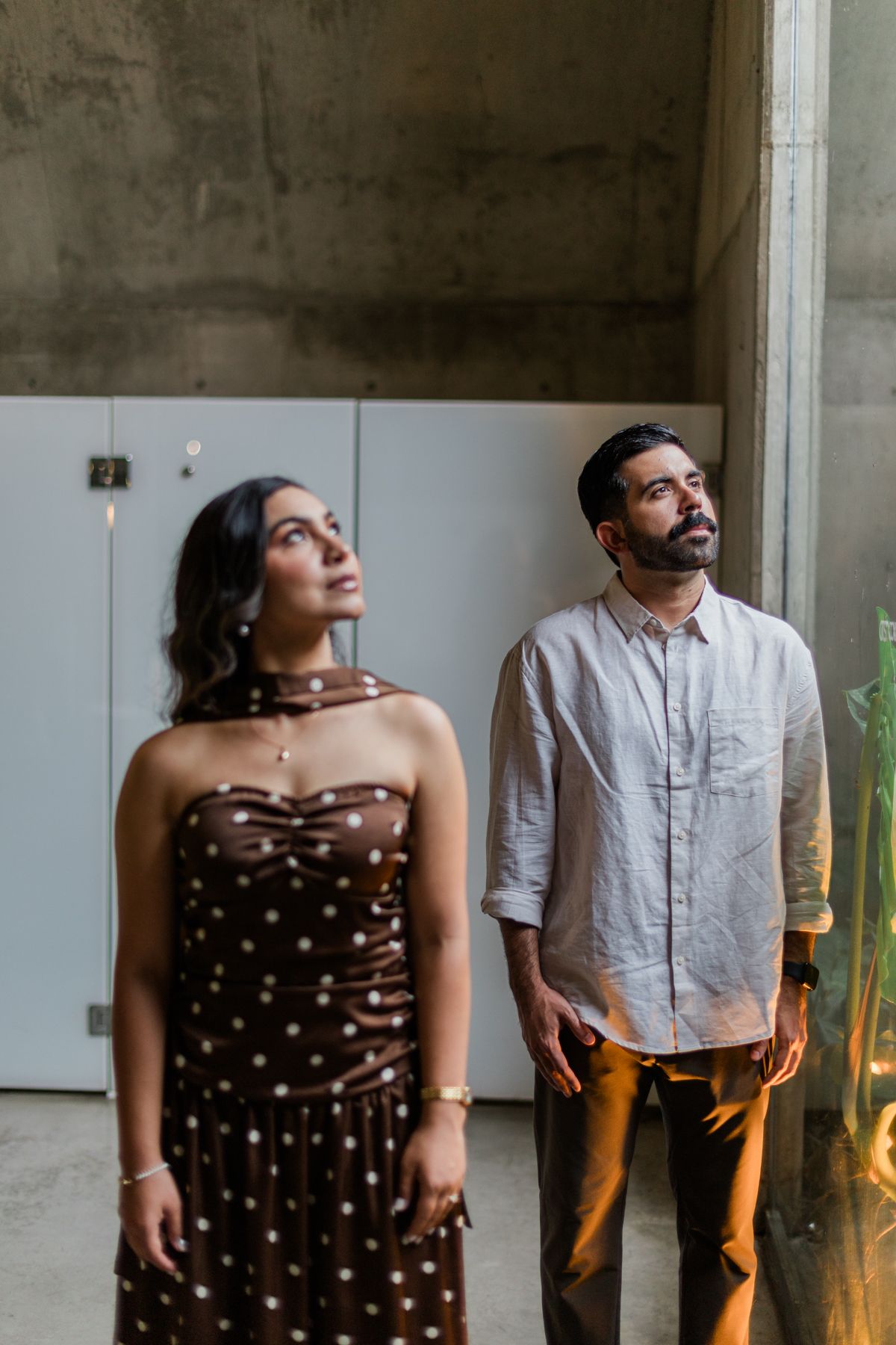 Couple gazing upward in concrete space, woman in brown polka dot strapless dress, man in linen shirt.