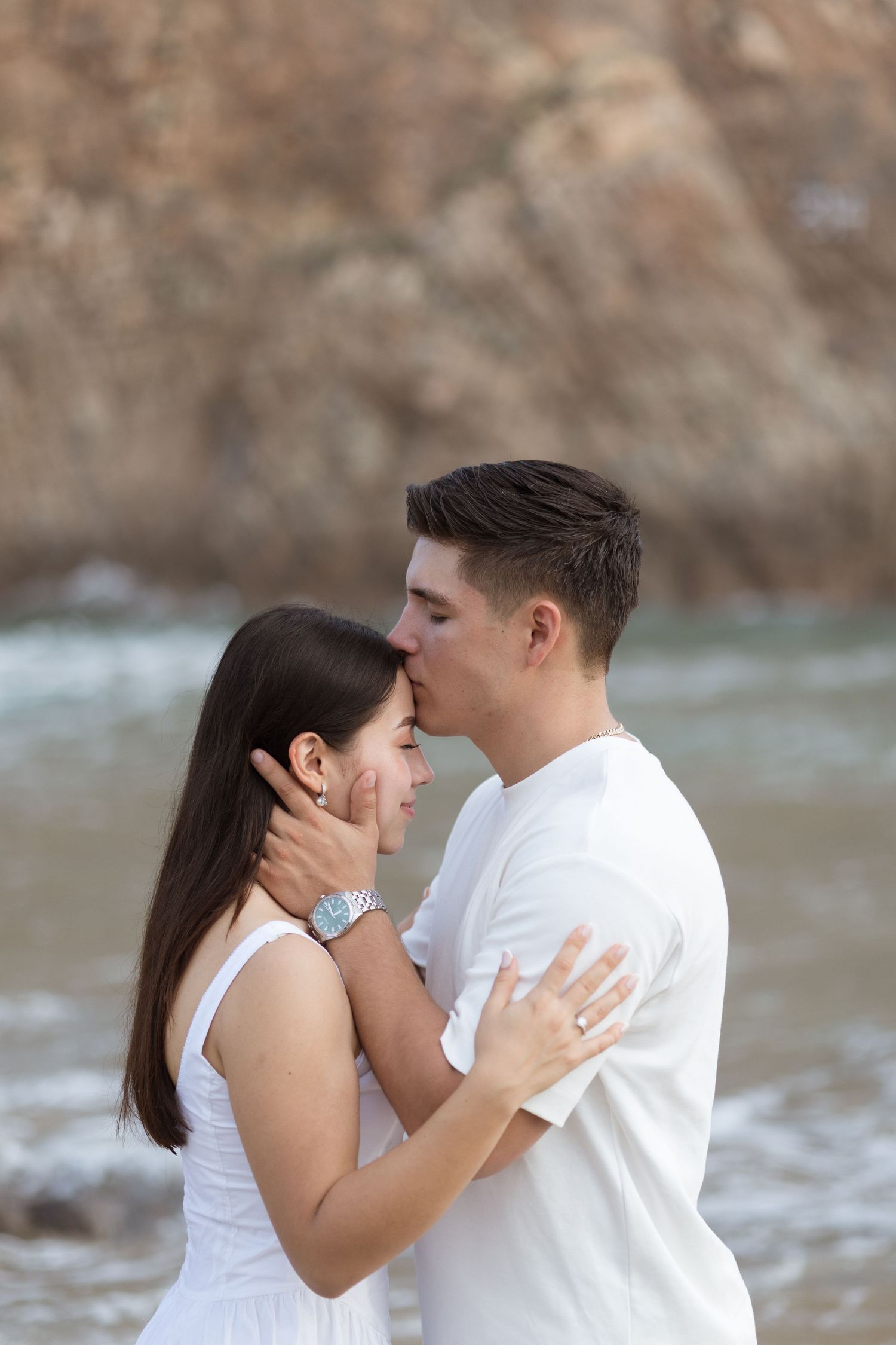 Romantic couple embracing and kissing on a beach, wearing white outfits with rocky cliffs in the background.