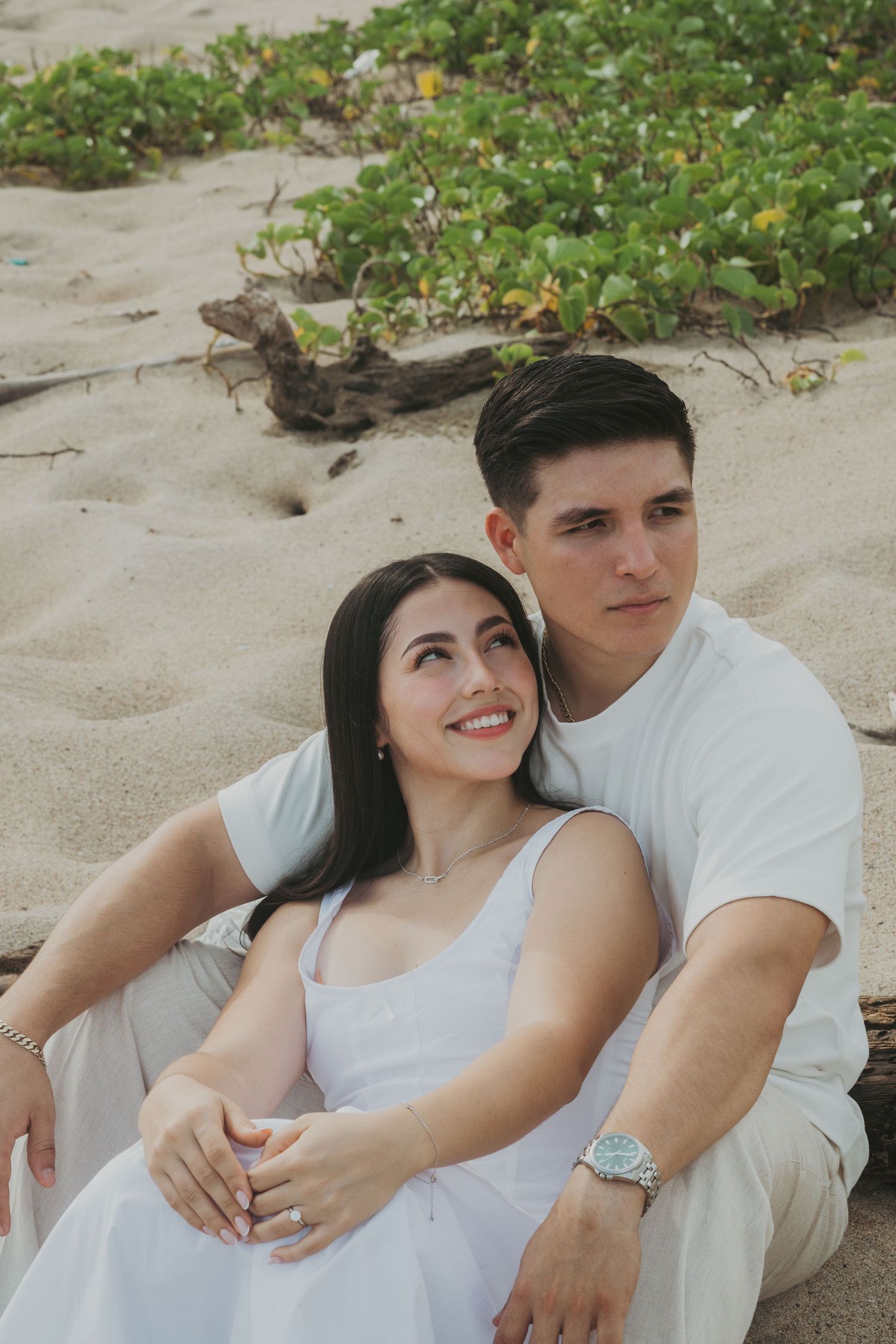 A couple in white outfits sits together on sandy beach, woman smiling, man holding her close with green plants behind.