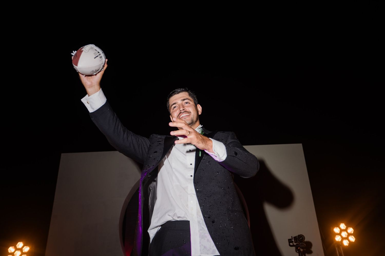 Dapper groom in black tuxedo proudly holds a white ball aloft while smiling on a lit outdoor wedding stage.
