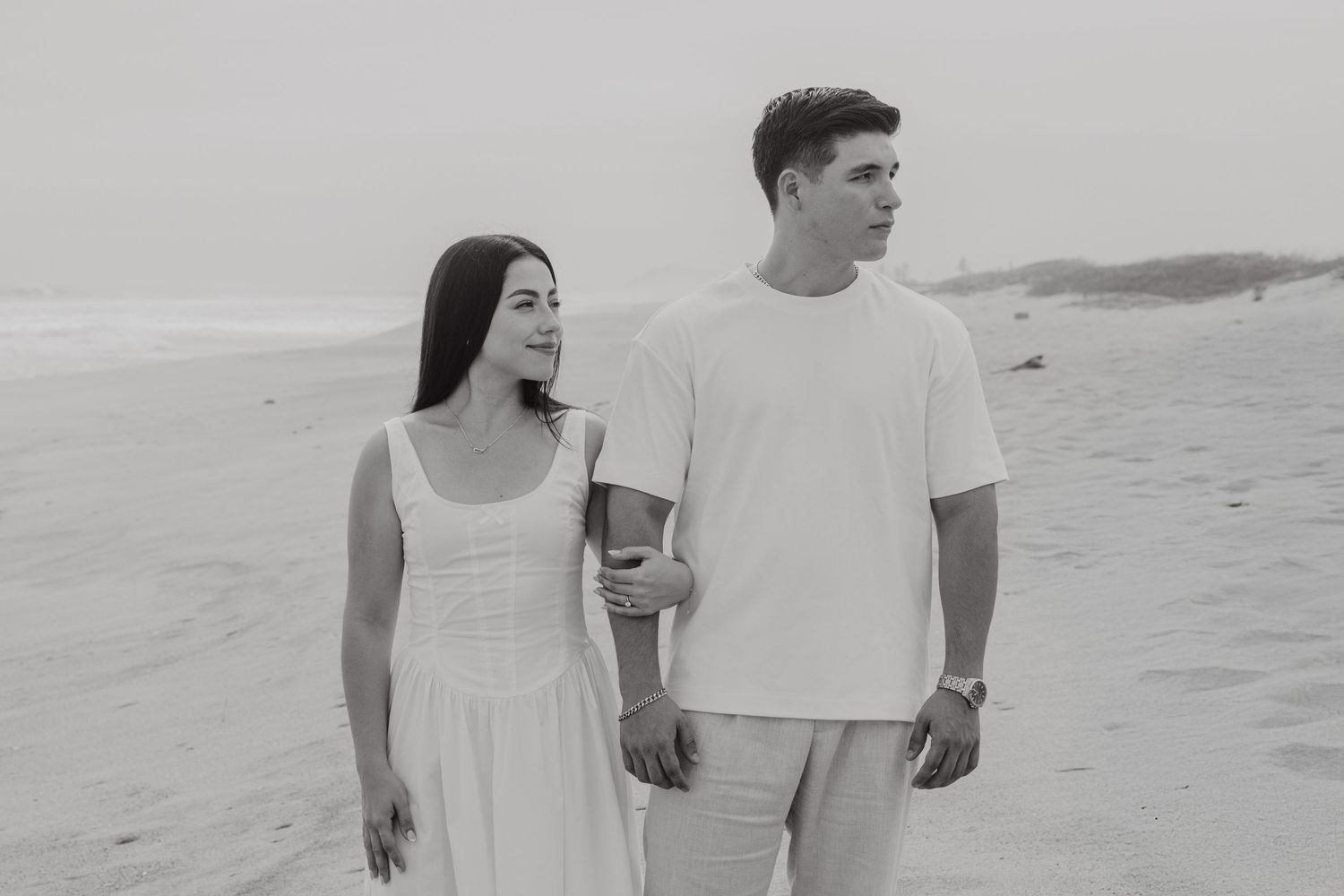 Black and white photo of a couple walking on a sandy beach, woman in white dress, man in white t-shirt.