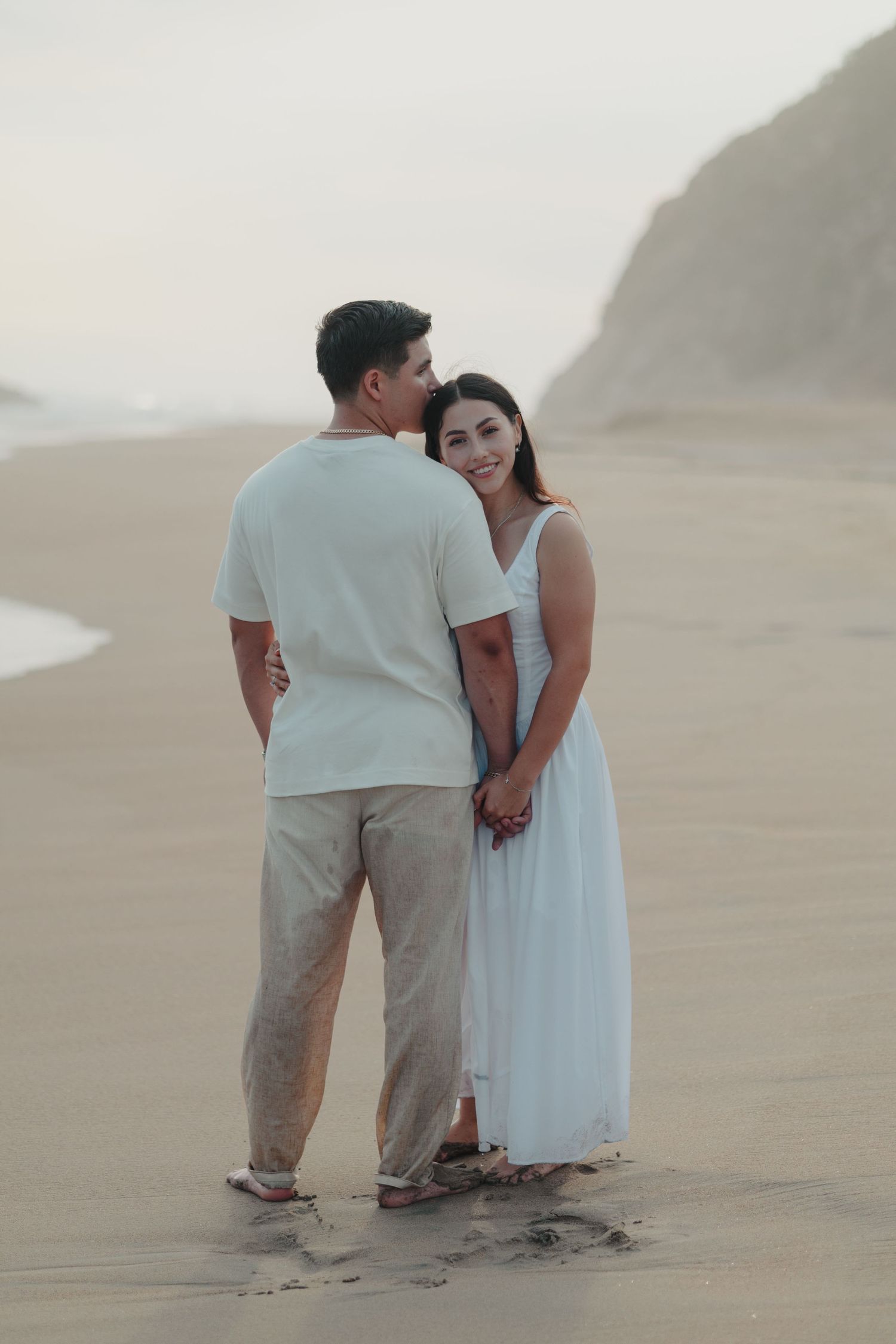 Couple holding hands on a sandy beach at dusk, woman in white dress smiling, man in white shirt and khaki pants.
