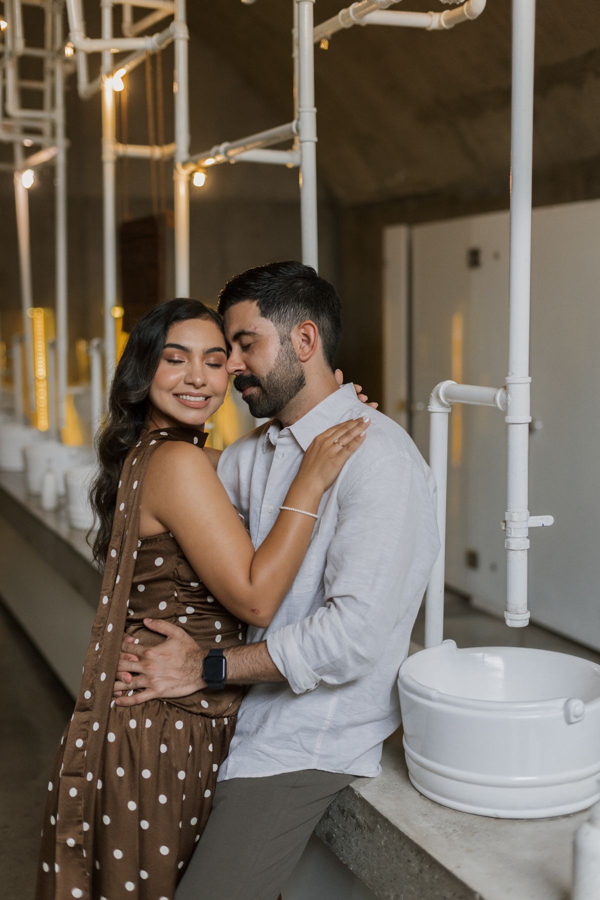 Romantic couple embracing near industrial pipes and string lights, woman in brown polka dot dress.