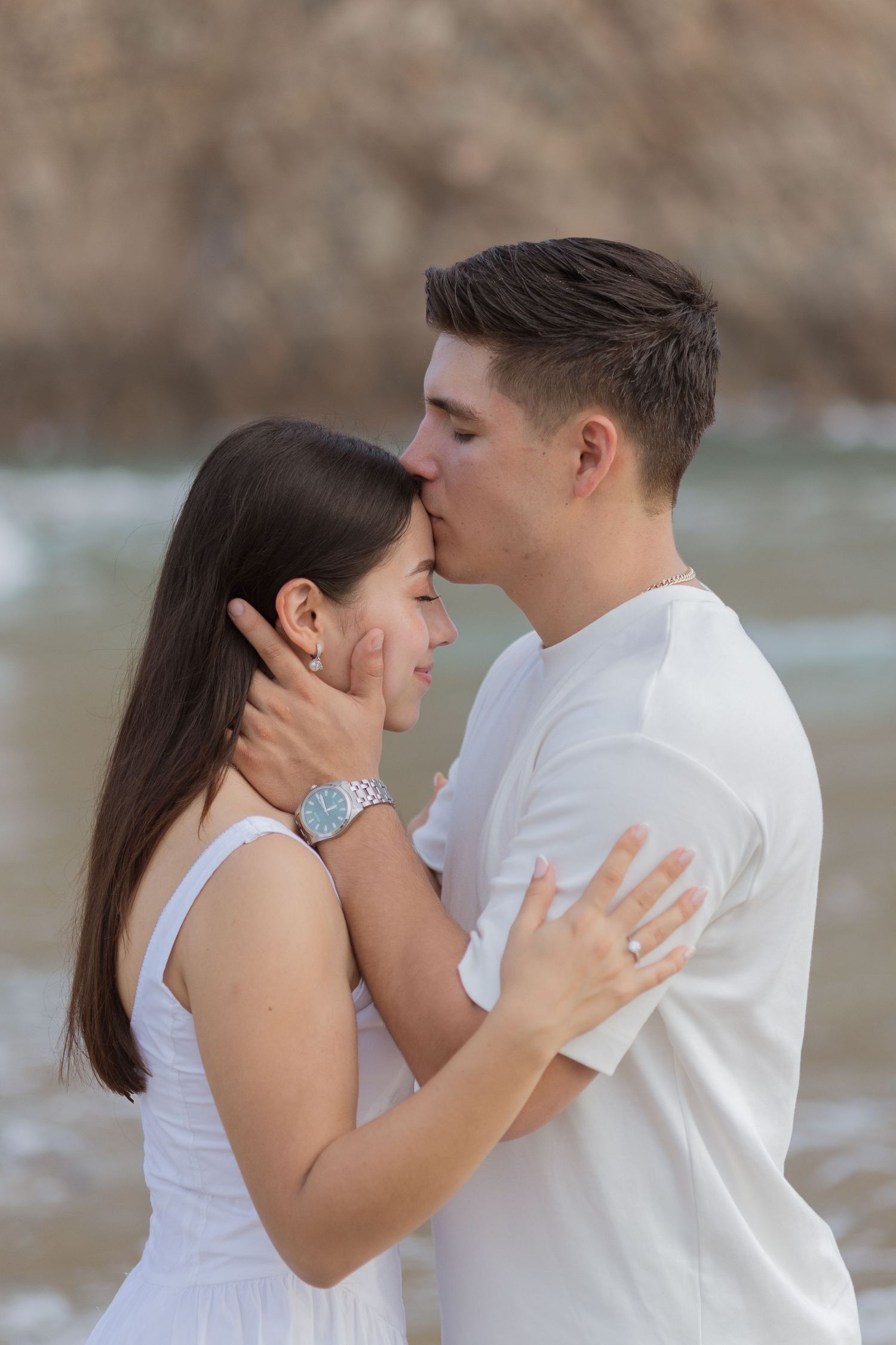 Young couple sharing a tender kiss on a rocky beach, dressed in white outfits near ocean waves.