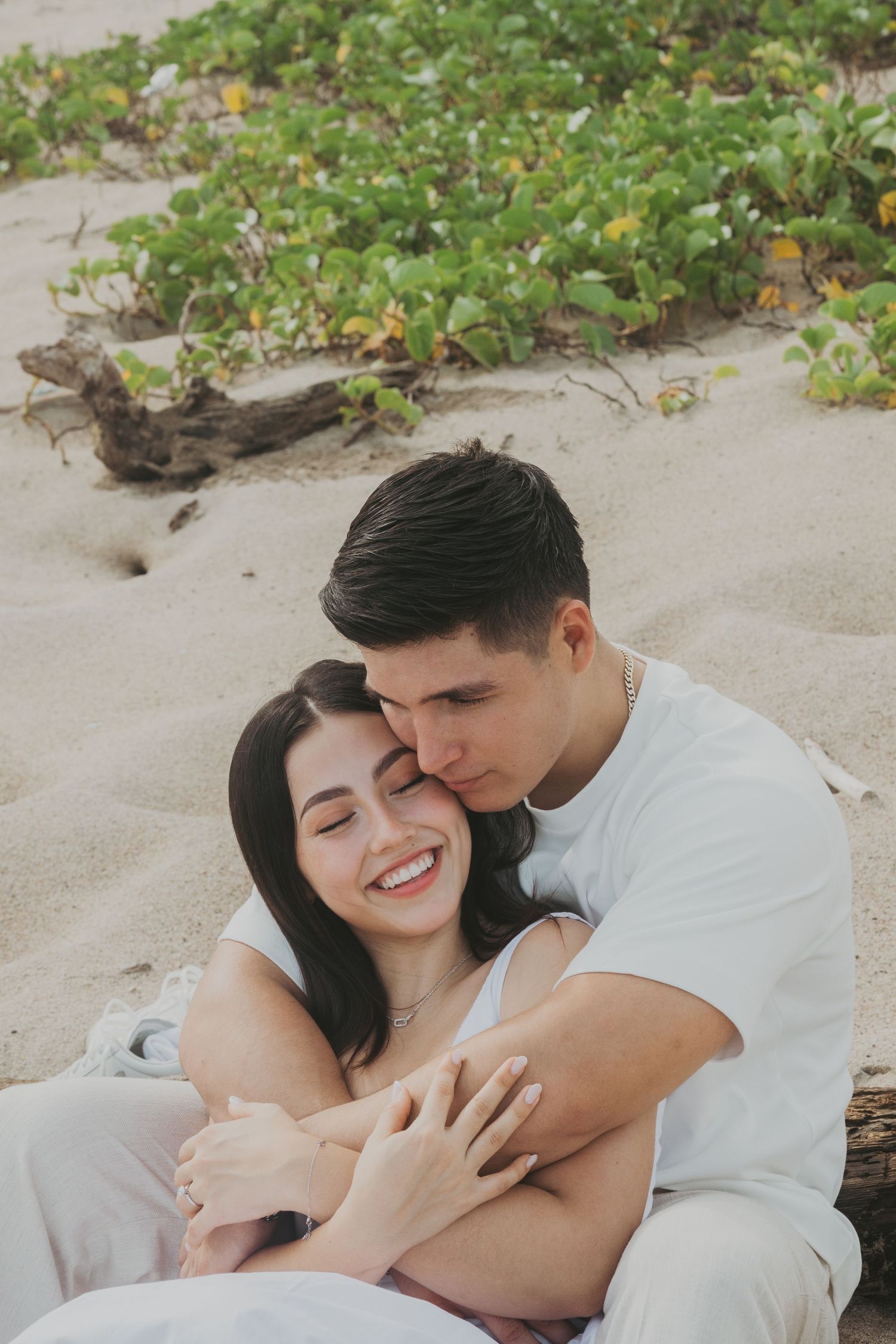 A couple embraces on a sandy beach, the woman smiling joyfully as the man kisses her cheek near lush green foliage.
