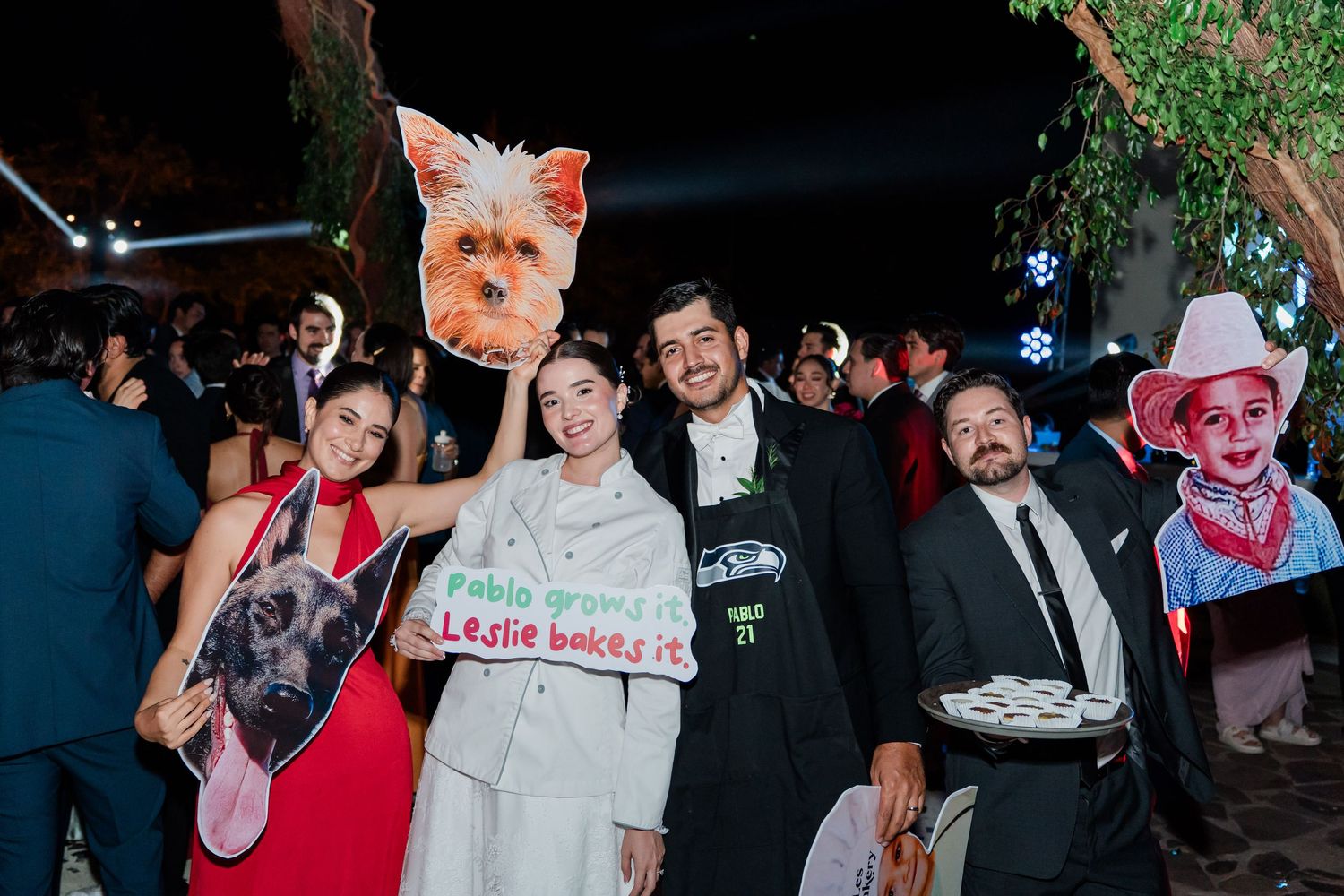 Wedding guests pose with fun photo props and signs at a lively outdoor evening wedding celebration.