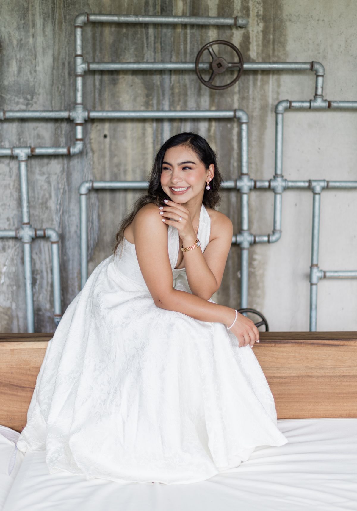 Smiling bride in white ball gown sitting on a wooden bed frame against an industrial concrete wall.