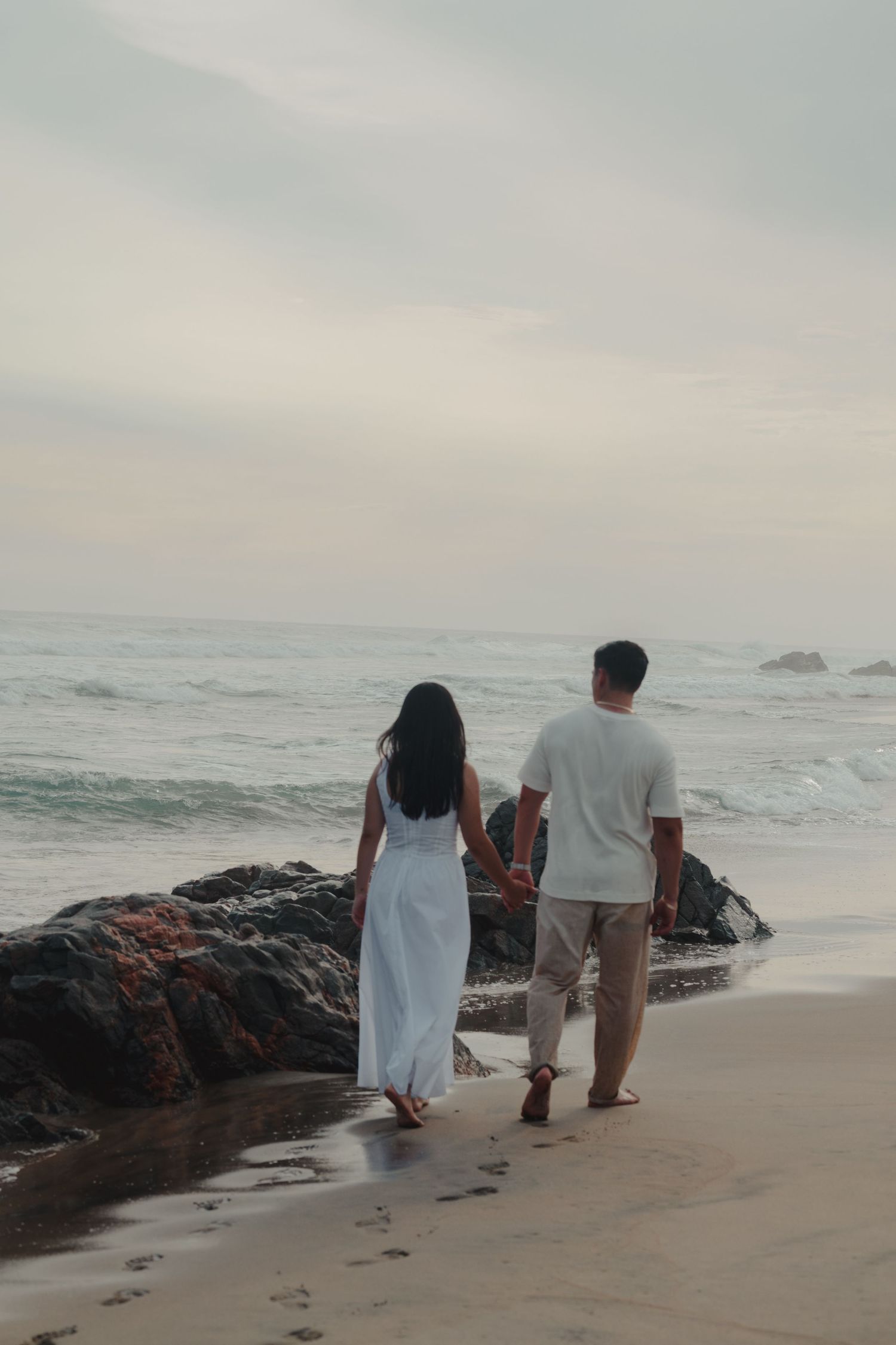 Couple holding hands walking on a sandy beach near rocks with ocean waves in the background at dusk.