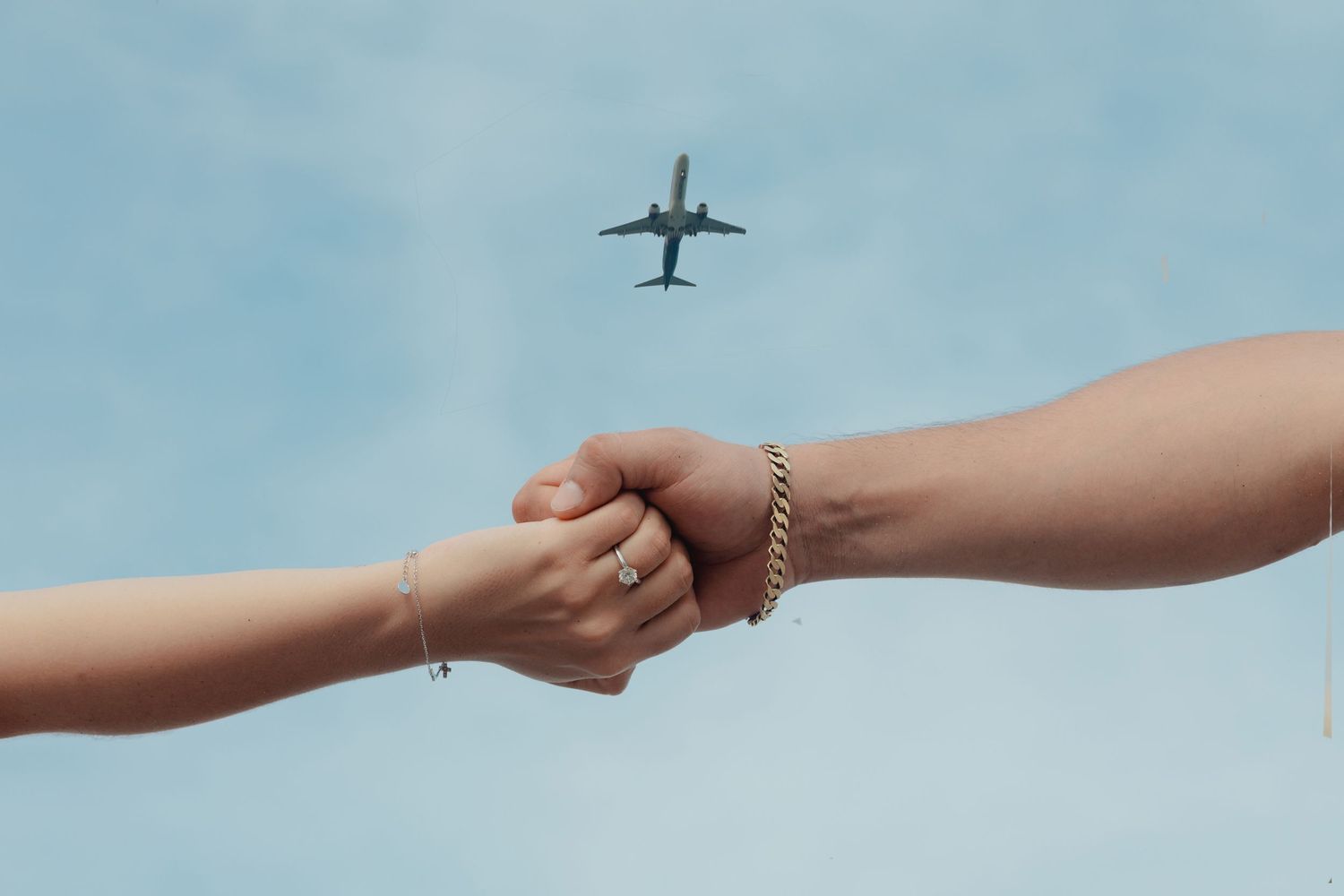 Two hands clasping together with a gold bracelet and ring, airplane flying overhead against a blue sky.
