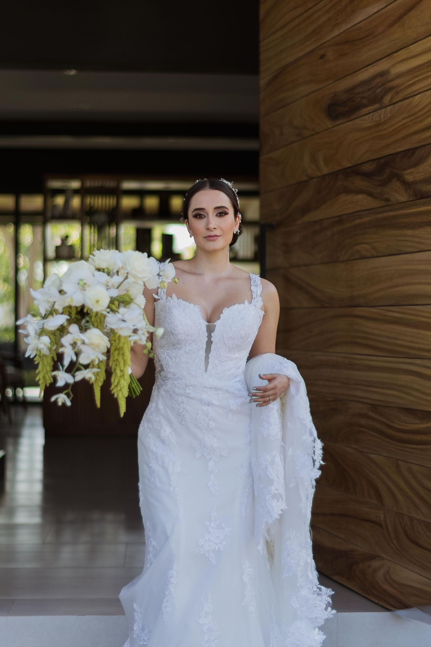 Stunning bride in fitted lace wedding dress with white floral bouquet posing by wooden wall.