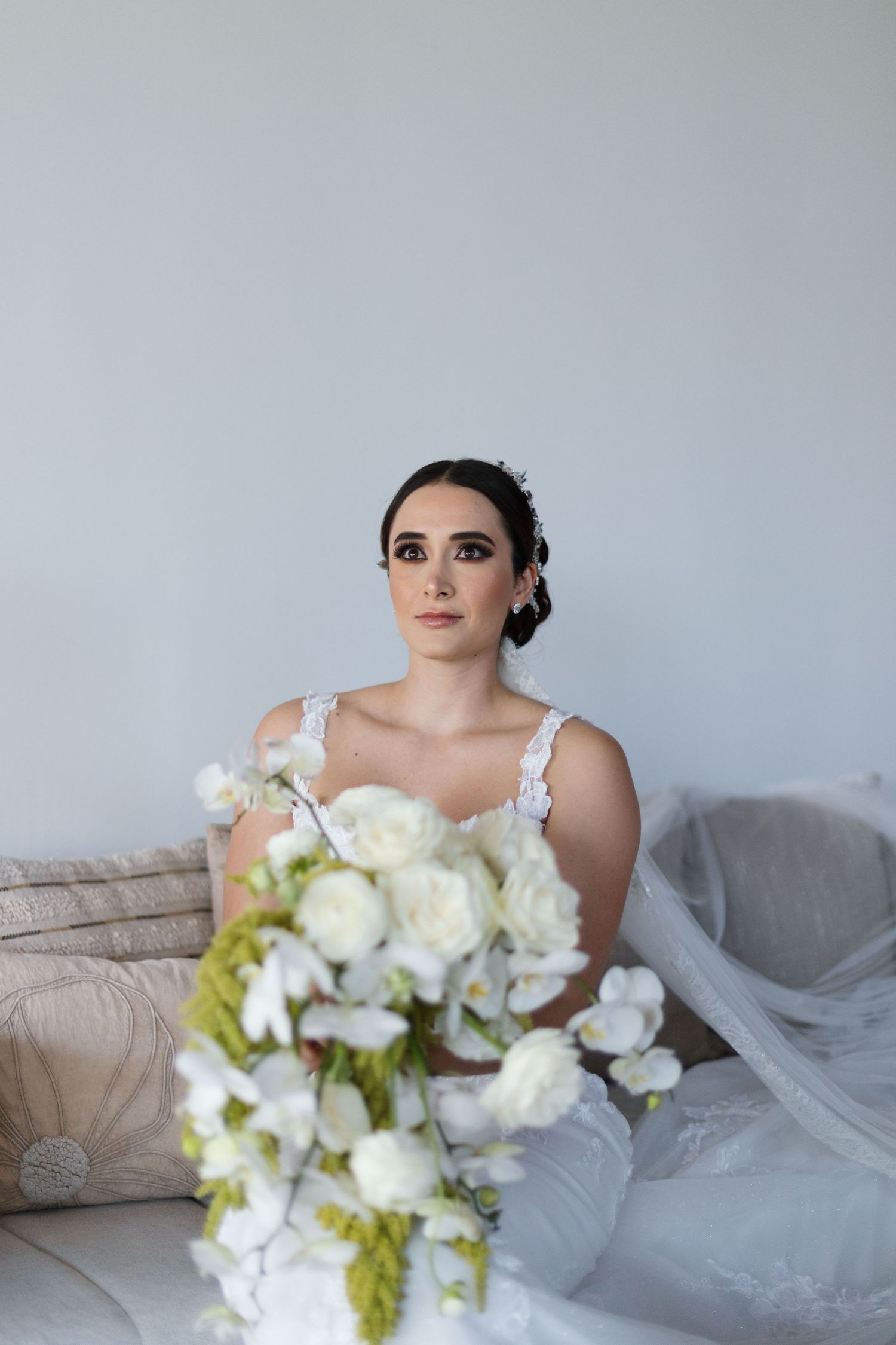 Elegant bride in lace gown seated on couch with cascading white orchid bouquet in foreground.
