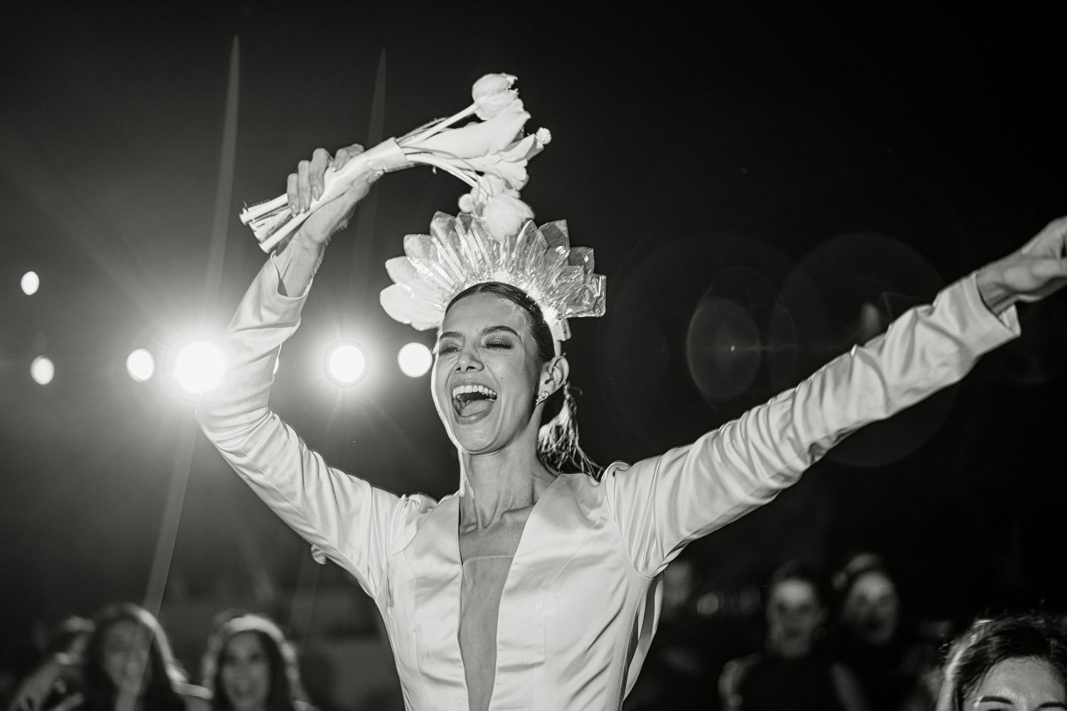 Bride in white outfit raises arms triumphantly wearing decorative crown headpiece at wedding celebration.