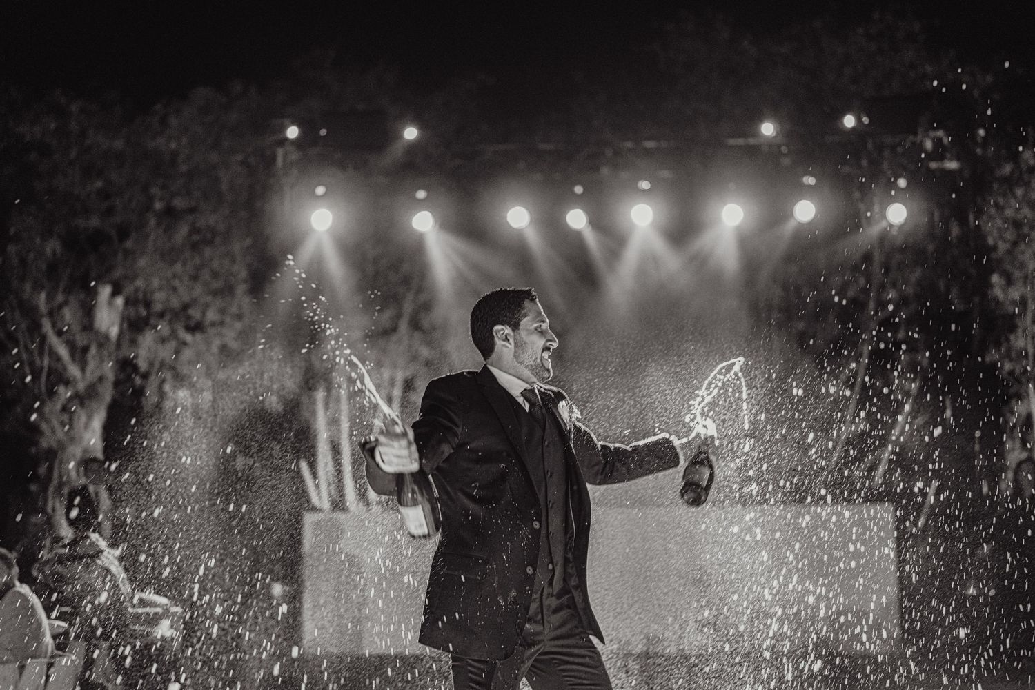Groom spraying champagne bottles under stage lights at dramatic black and white wedding celebration.