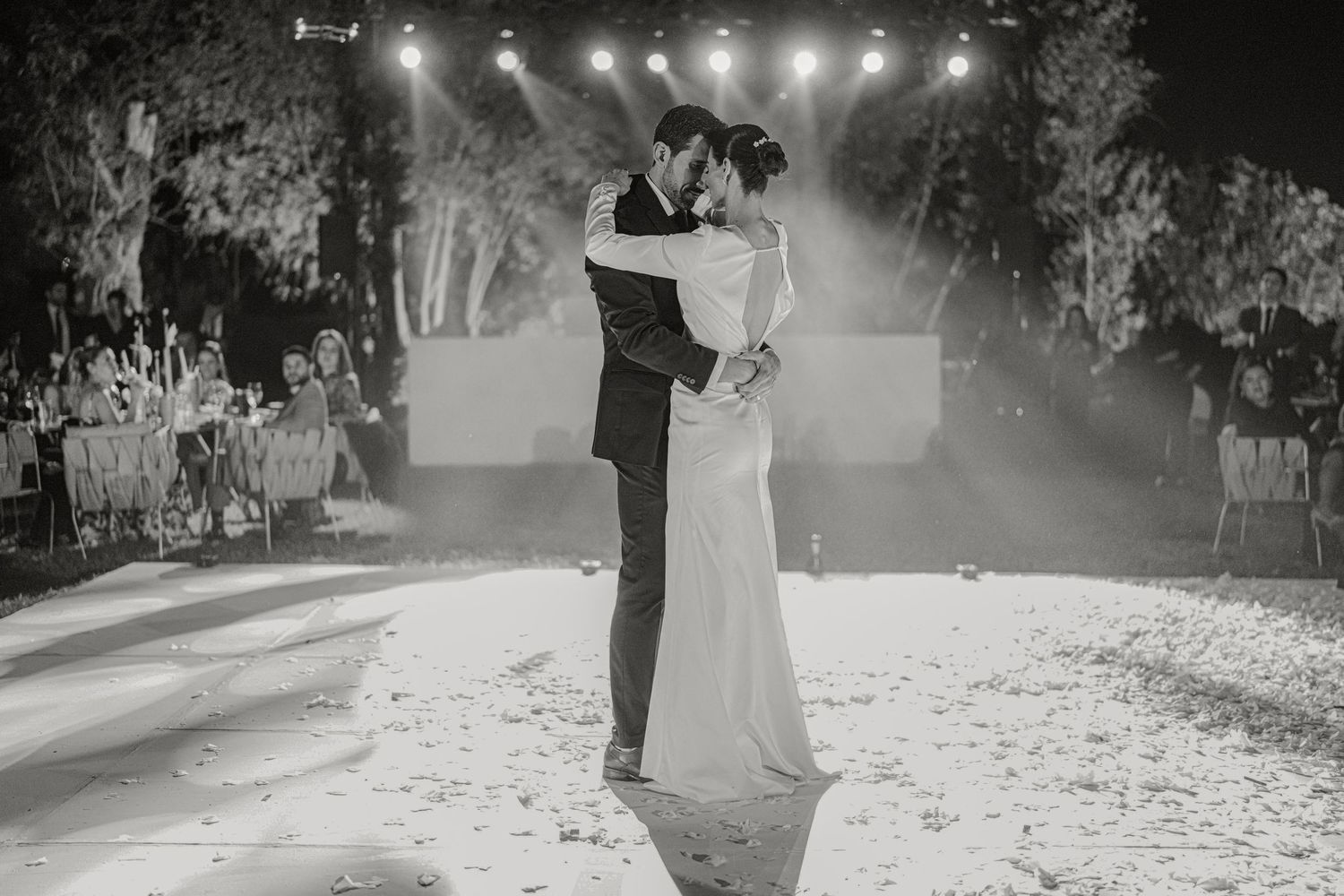 Bride and groom sharing first dance under spotlights at elegant outdoor wedding in black and white.