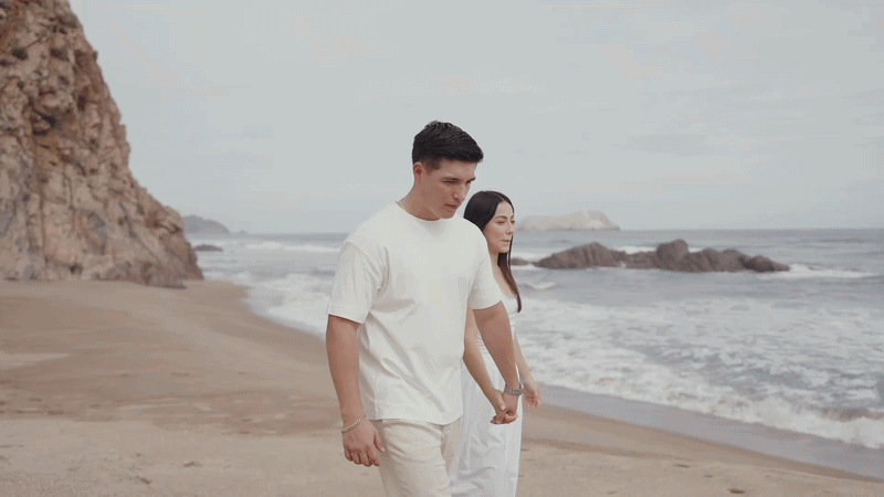 A couple walks hand in hand on a sandy beach near rocky cliffs, dressed in white outfits with ocean waves behind them.