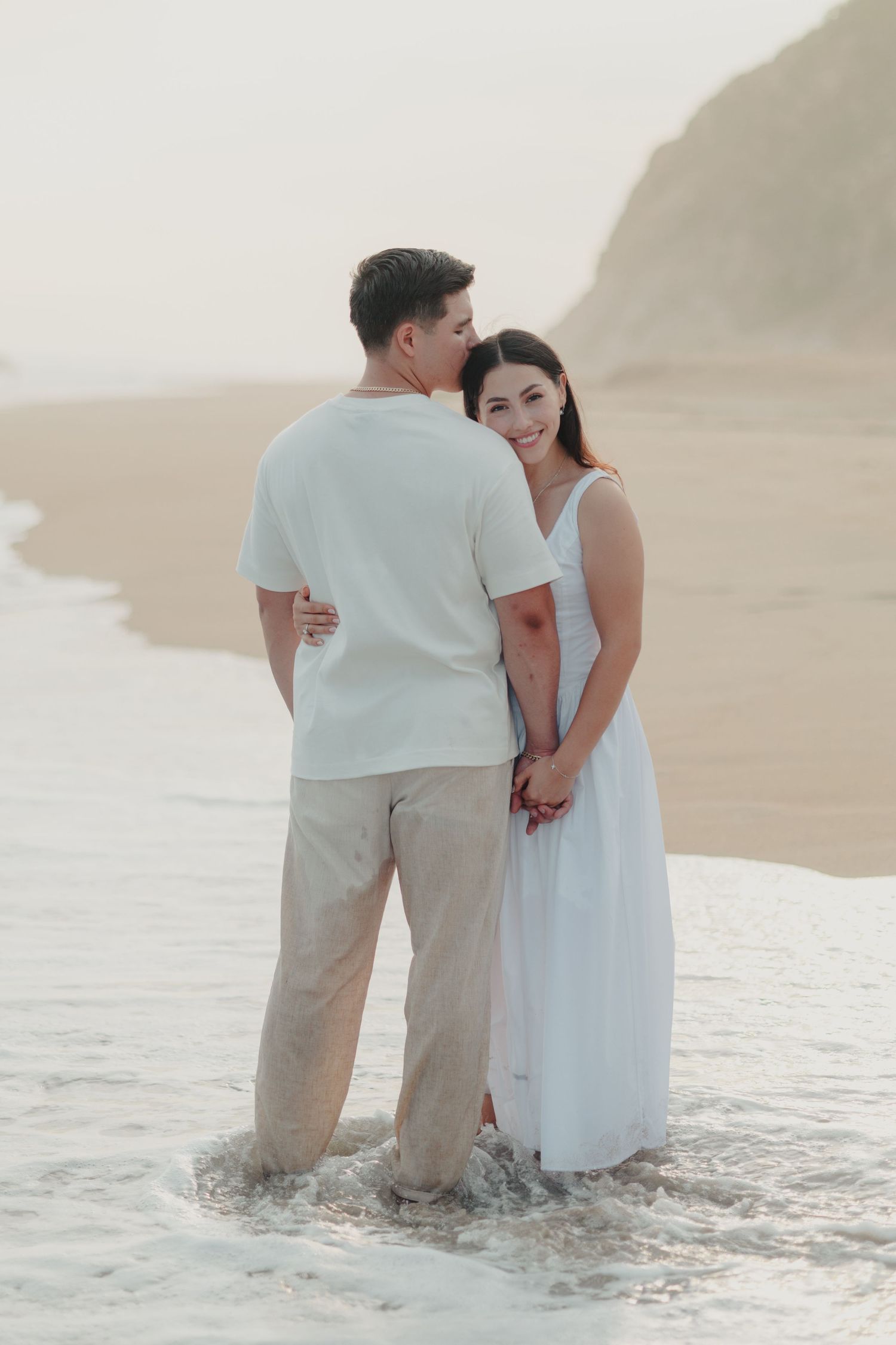 Couple standing in ocean waves at beach, woman in white dress smiling, man in white shirt and khaki pants.