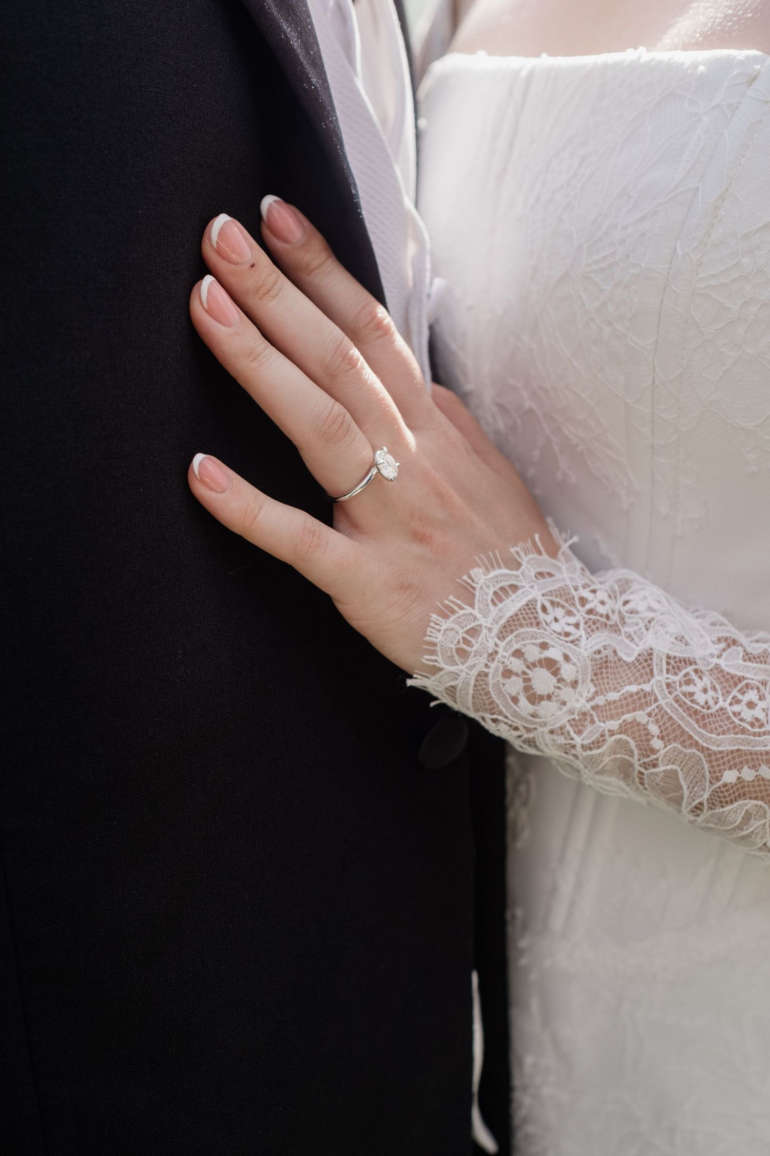 Bride in lace wedding dress placing hand on groom's black suit jacket showing diamond engagement ring.