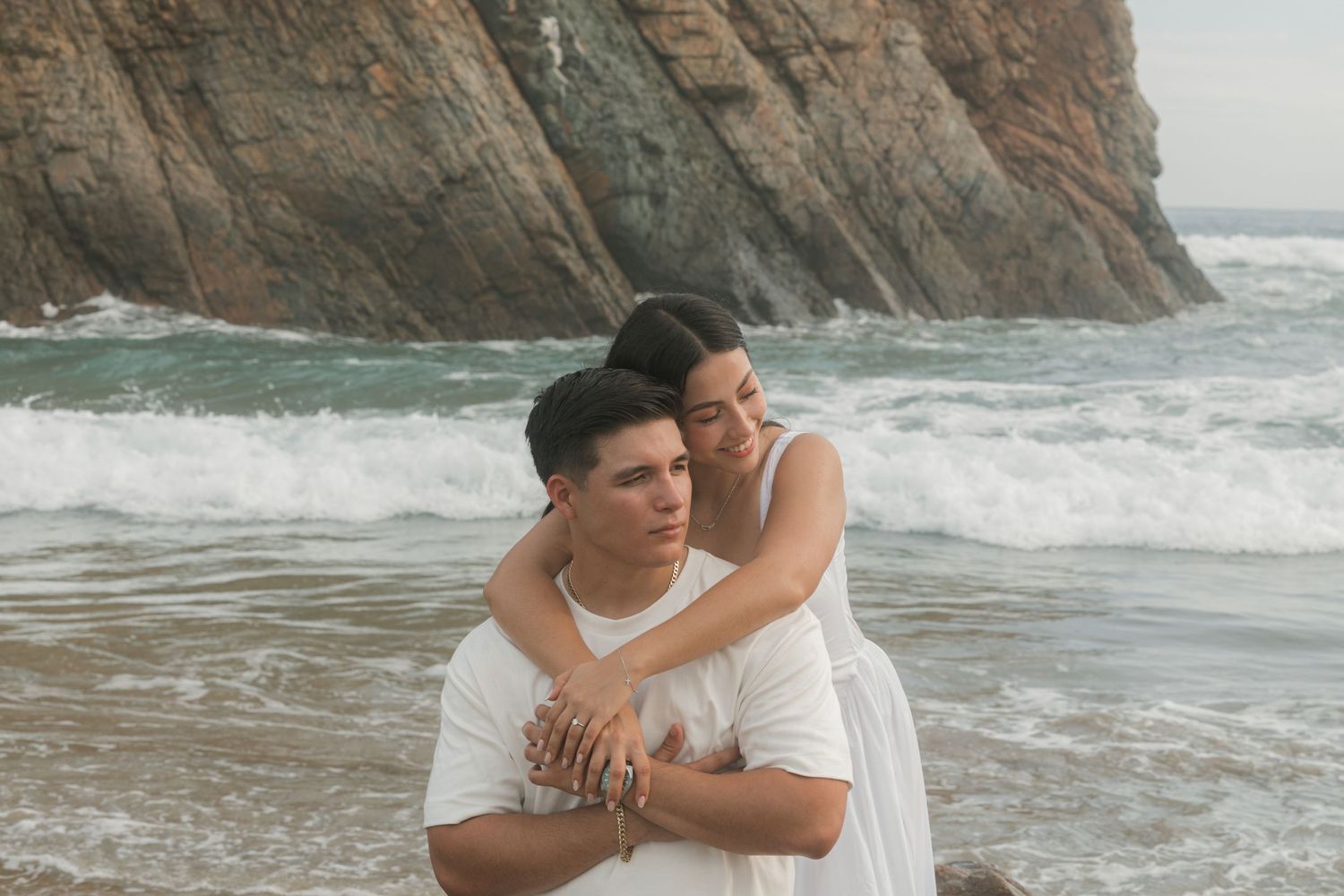 Couple embracing on a rocky beach, wearing white outfits with ocean waves and dramatic cliffs in the background.