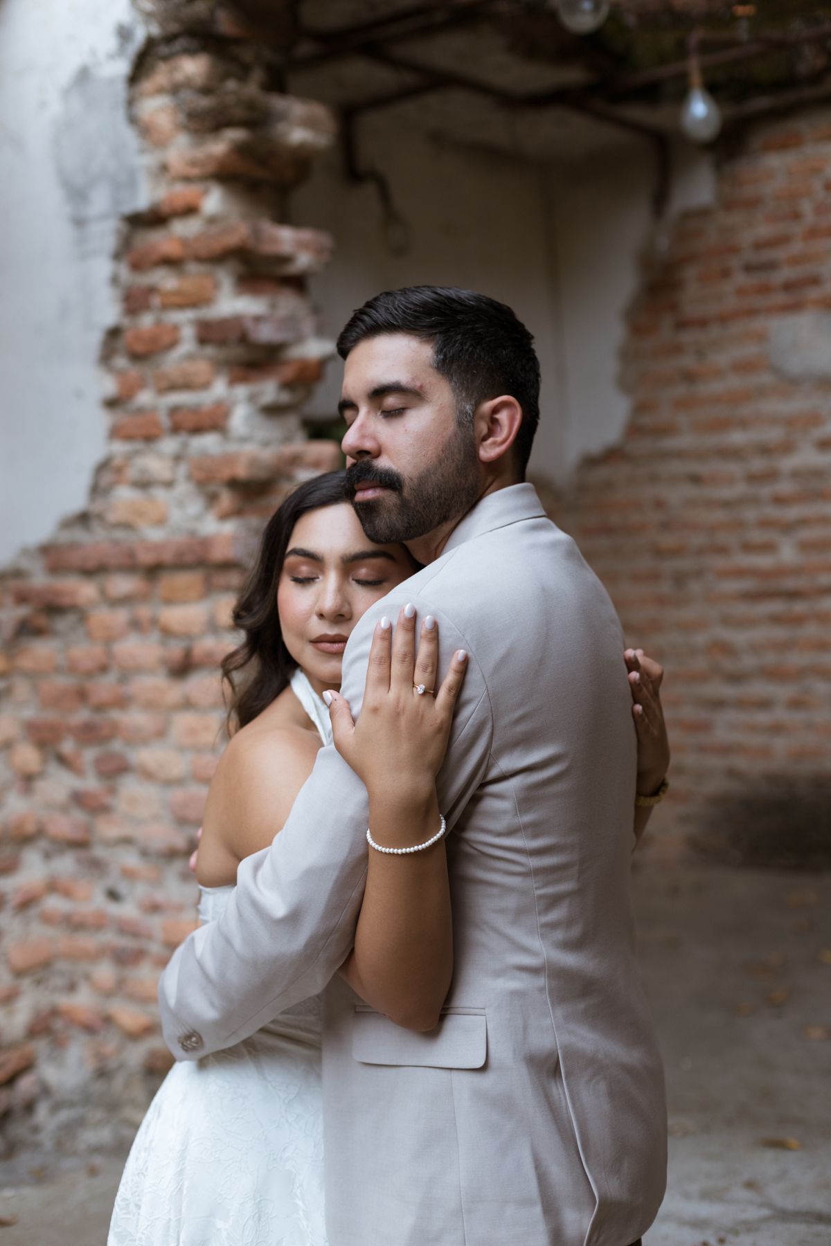 Bride and groom share tender embrace near exposed brick wall, both with eyes closed peacefully.