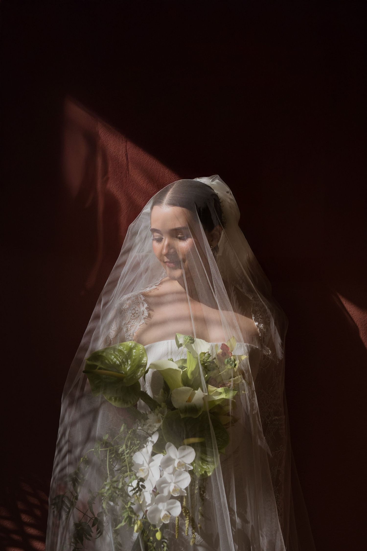 Elegant bride in white lace gown holding green and white floral bouquet under veil in dramatic lighting.