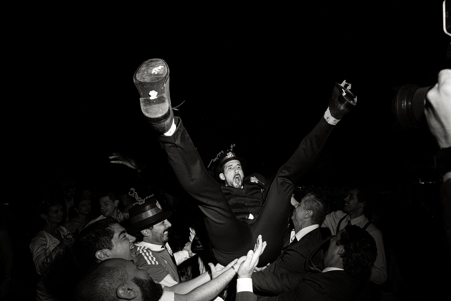 Groom lifted by cheering wedding guests in energetic black and white crowd celebration photo.