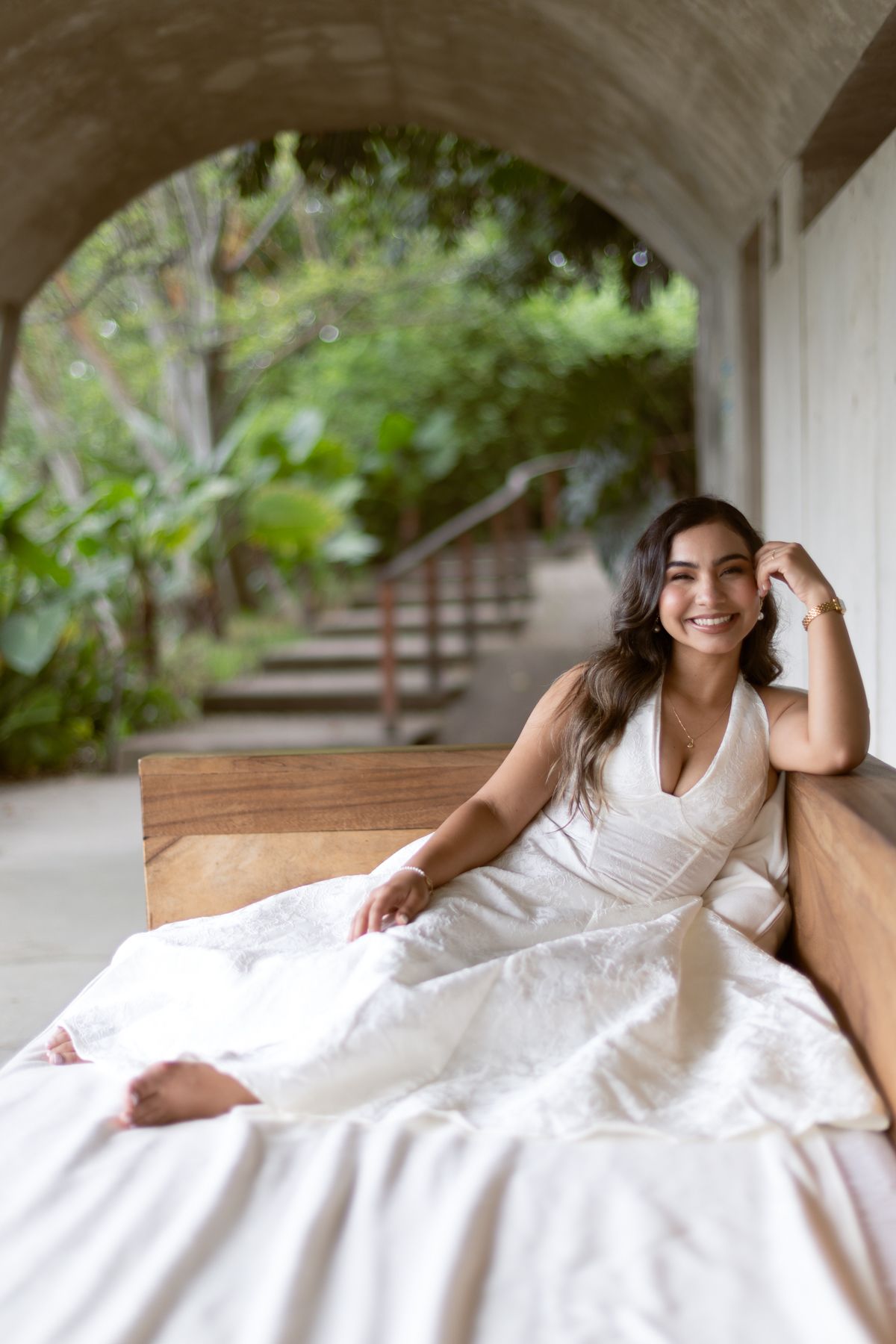 Smiling bride in white halter dress sitting barefoot on an outdoor daybed under a stone archway.