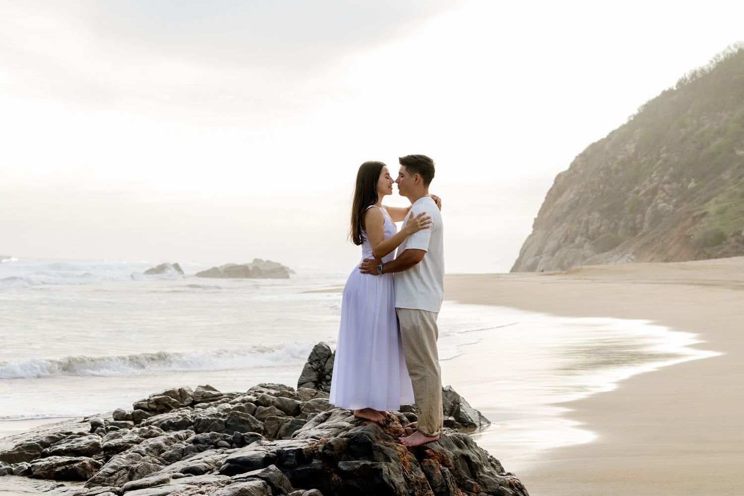 Romantic couple kissing on rocks at a scenic beach with ocean waves and green cliffs in the background.