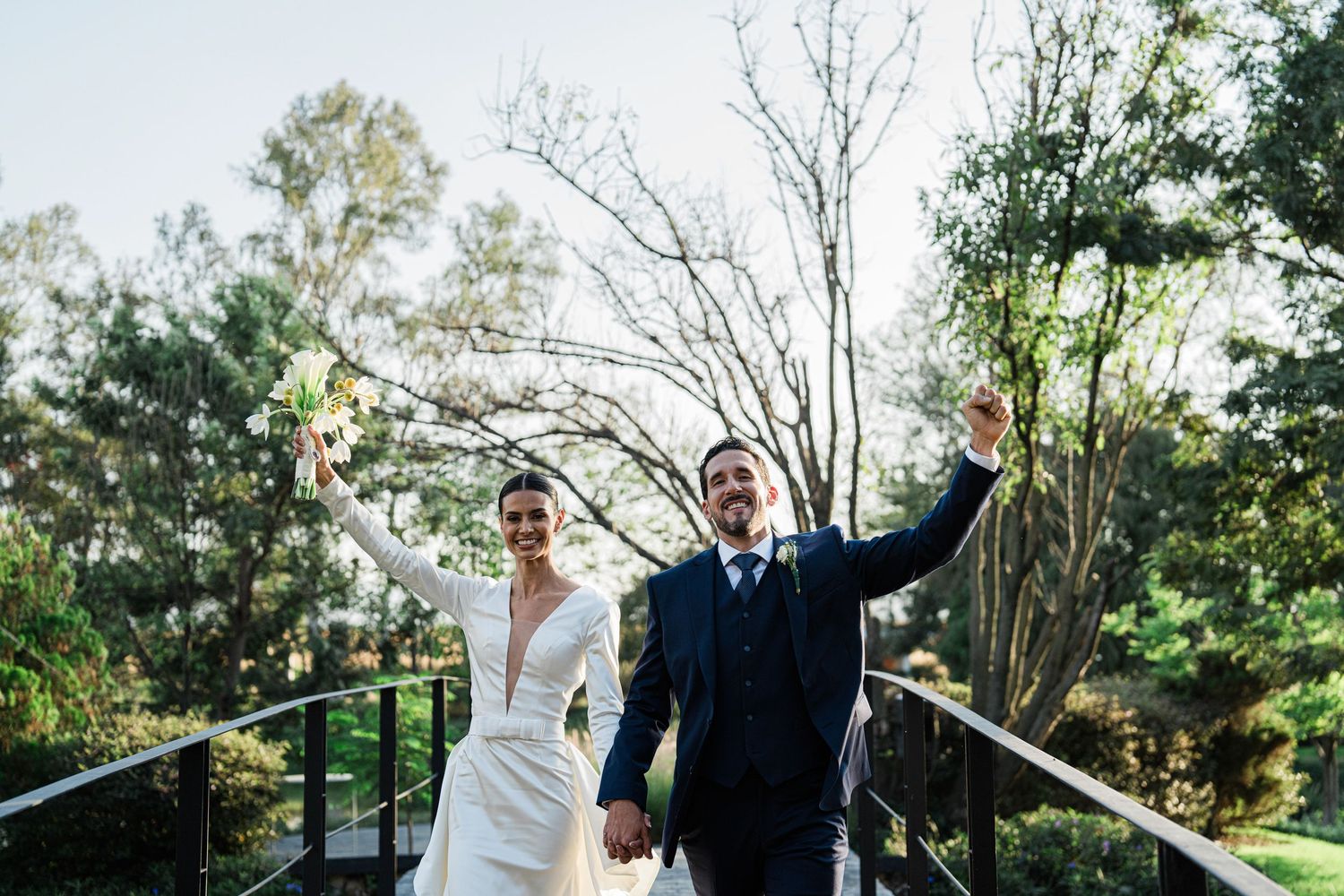 Joyful newlyweds raising hands in celebration on bridge surrounded by trees.