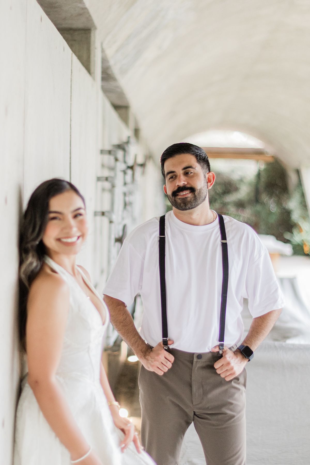 Smiling groom in white shirt and suspenders stands with bride in white dress in a bright corridor.