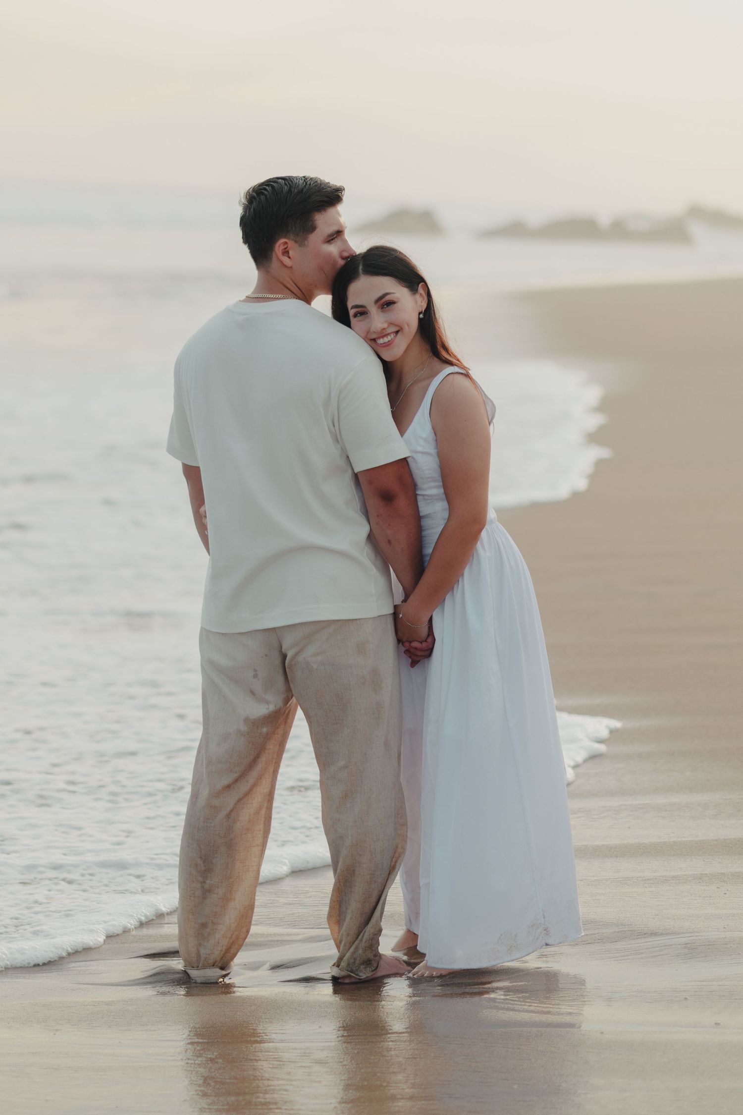 Couple holding hands on a beach at sunset, woman in white dress smiling, man in beige outfit near ocean waves.